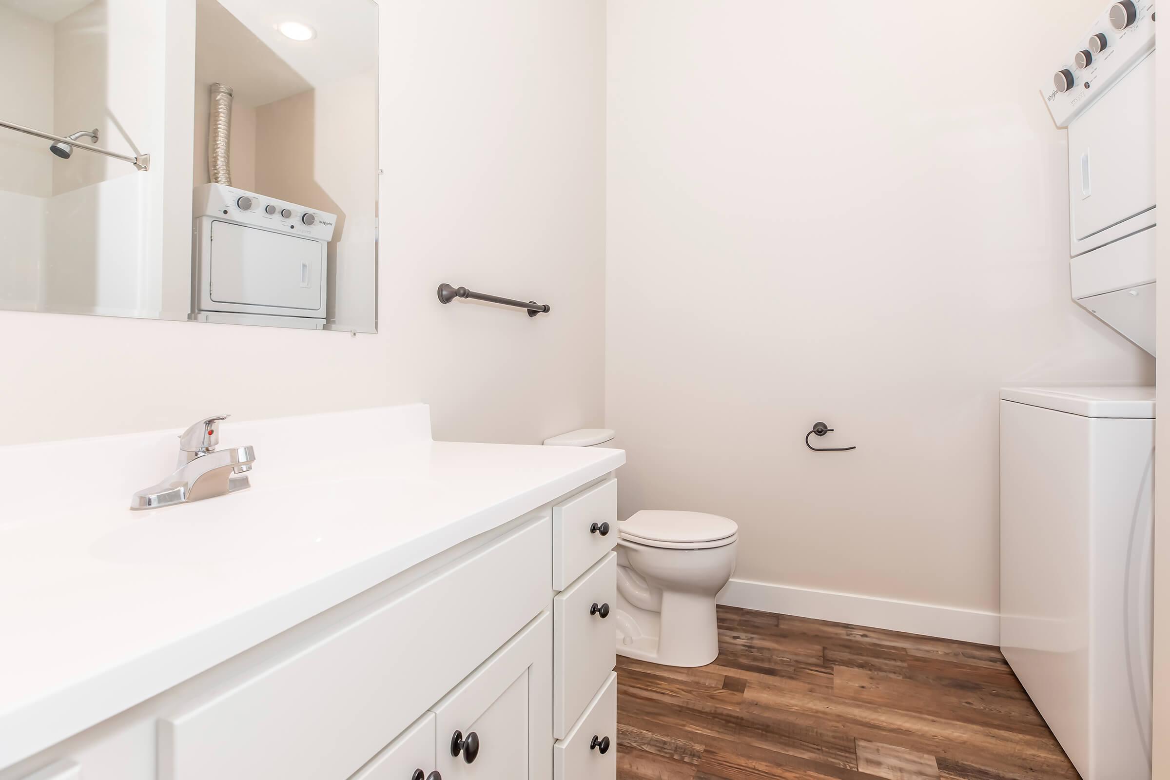 A clean and modern laundry room with a white sink and cabinetry, a washing machine and dryer stacked on one side, and a toilet on the opposite side. The room features light-colored walls and dark wood-style flooring, creating a spacious and airy atmosphere.