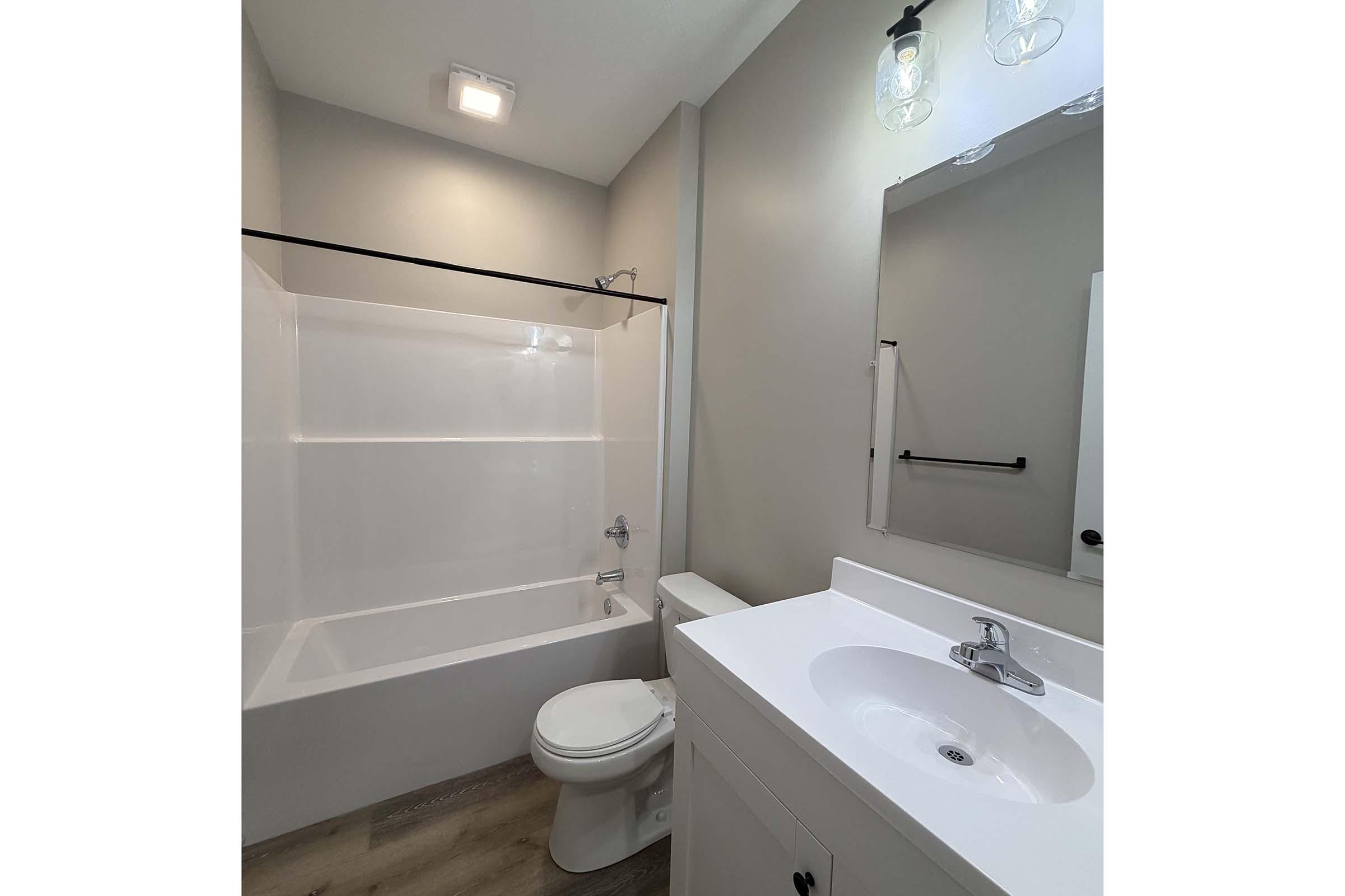 A modern bathroom featuring a white bathtub with a shower, a sleek vanity with a sink, a large mirror, and neutral-colored walls. The space is well-lit with a ceiling light and two pendant lights above the mirror, creating a bright and clean atmosphere.