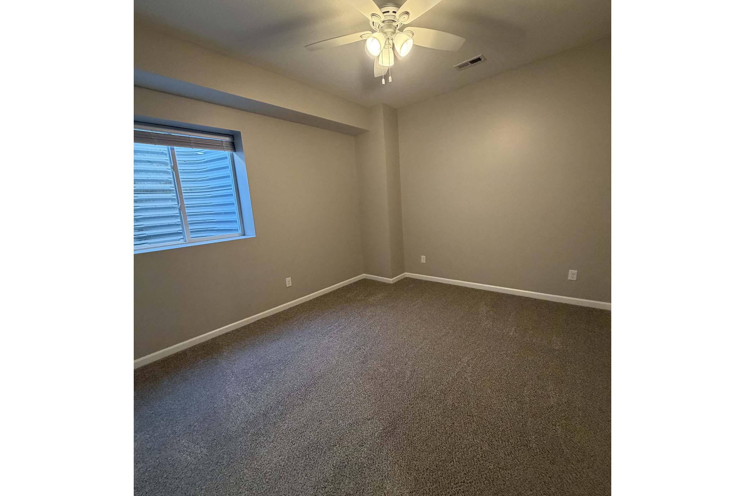 Empty room with beige walls and carpet. A ceiling fan hangs from the ceiling, and there is a window on the left side with light filtering through. The room is sparsely decorated with no furniture present, creating a spacious and clean appearance.