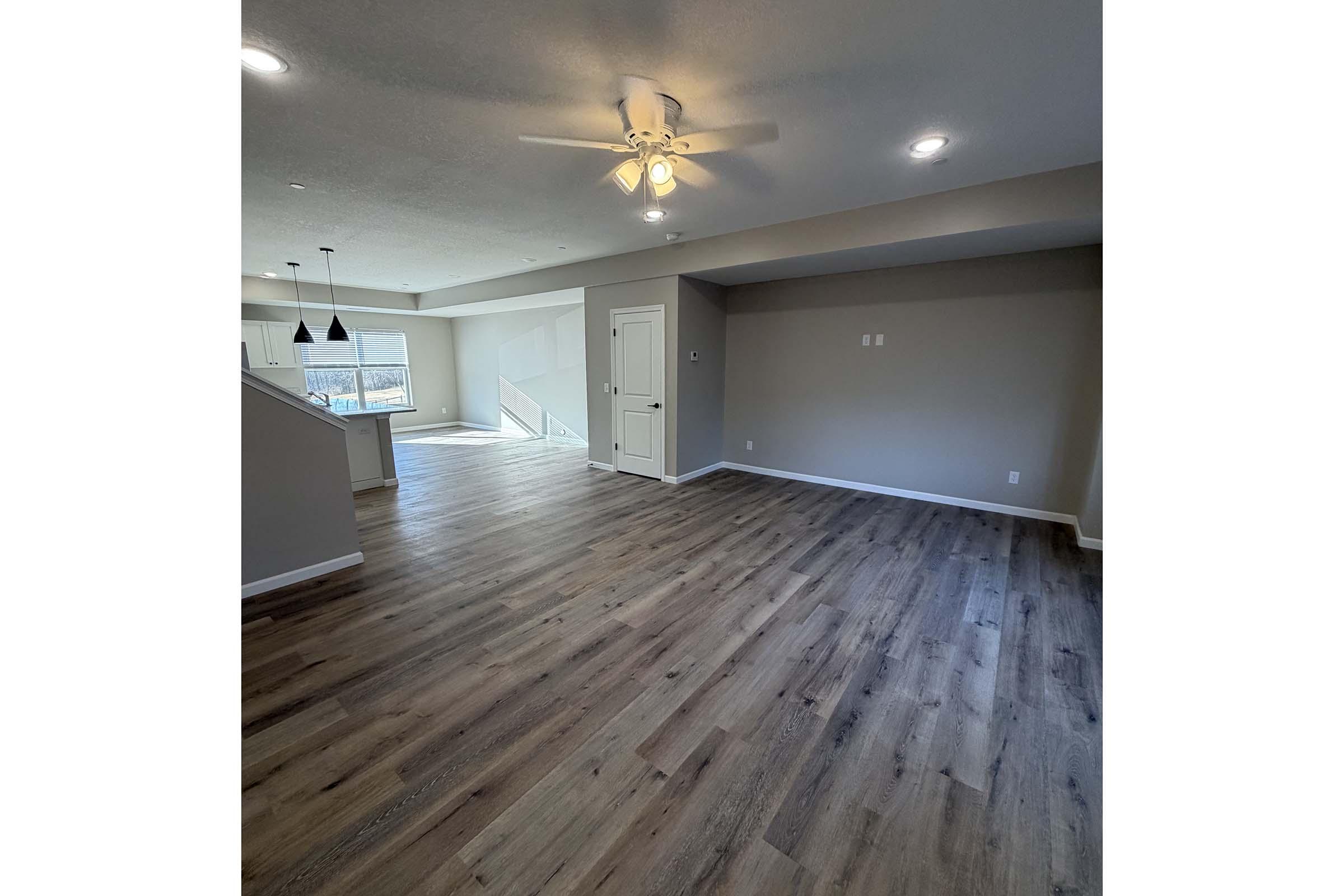 A spacious, well-lit interior room featuring wooden flooring, a ceiling fan, and neutral-colored walls. Windows allow natural light to flow in, and an open layout leads towards another room in the background. The space is clean and inviting, ideal for living or dining purposes.