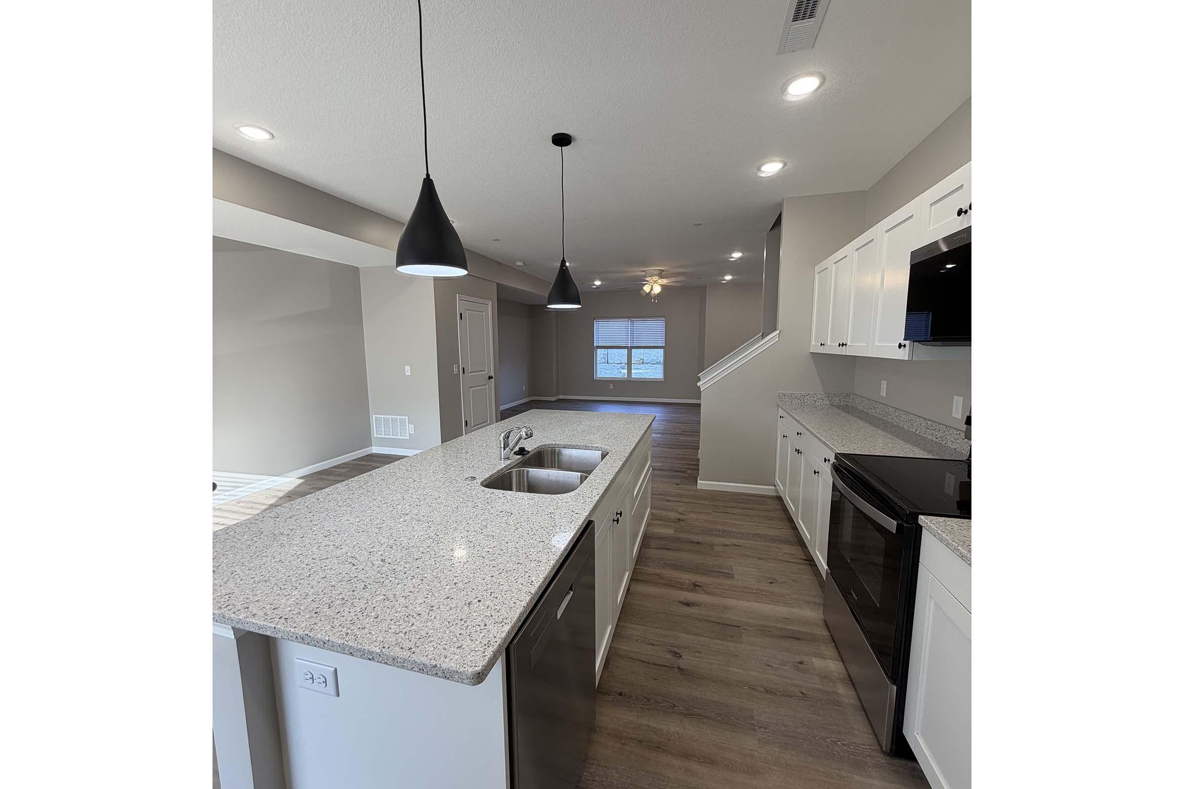 A modern kitchen featuring a large granite countertop with a double sink, white cabinets, and stainless steel appliances. Pendant lights hang above the counter, and the open concept design leads into a spacious living area with a ceiling fan and large windows. The flooring is a light wood finish.