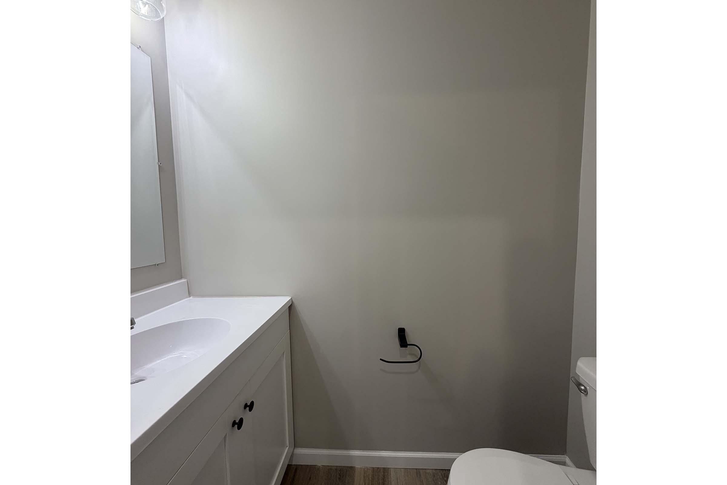 A clean, simple bathroom featuring a light-colored wall, a white vanity with a countertop sink, a large mirror above the sink, and a toilet. The flooring appears to be wooden. There is a wall-mounted electrical outlet near the floor, and the overall atmosphere is bright and uncluttered.