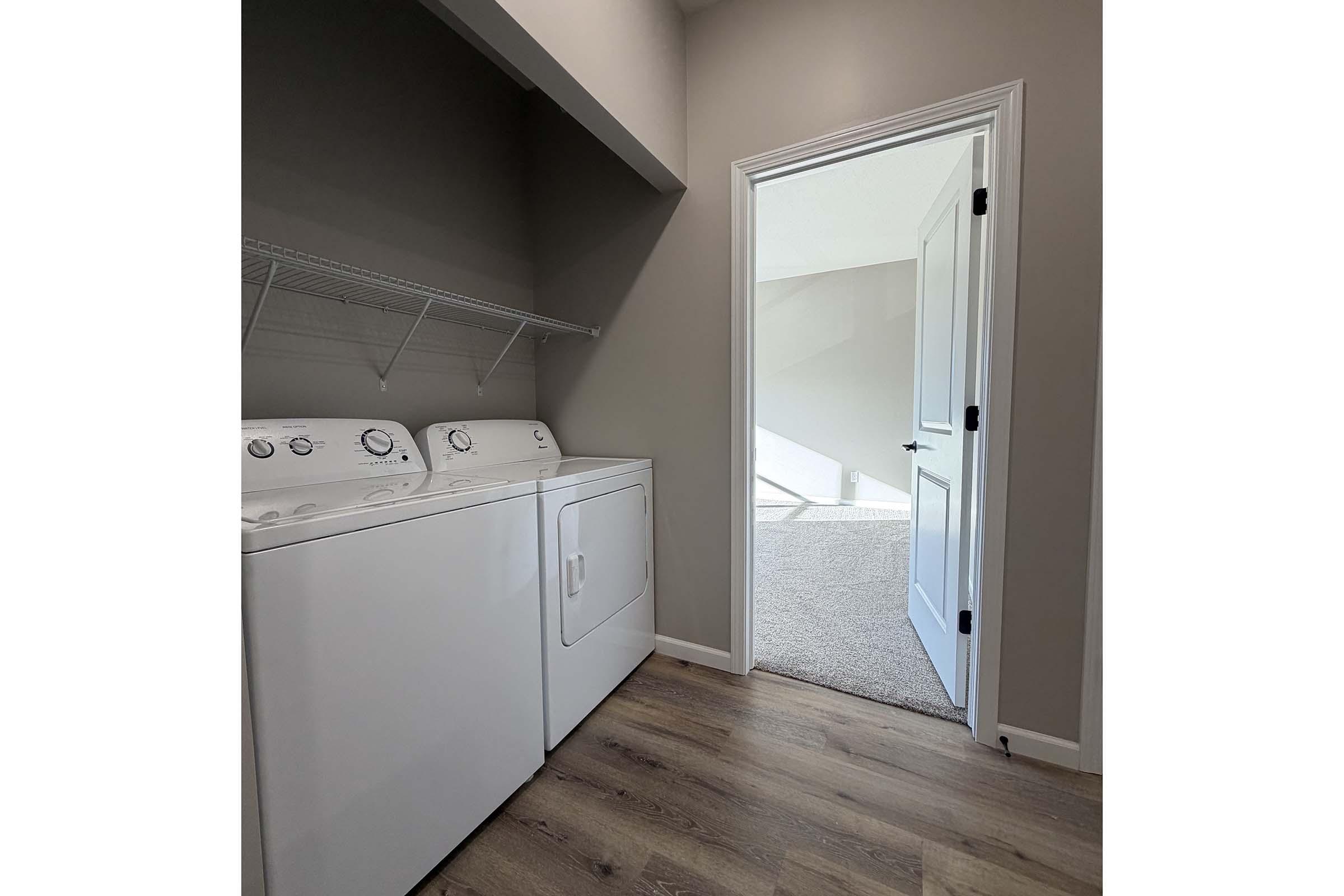 A laundry room featuring a white washer and dryer, with a wall-mounted shelf above. A light-colored door leads to another room, and the floor is made of wood. The walls are painted in a neutral tone, creating a clean and modern aesthetic. Natural light enters from the adjacent room.