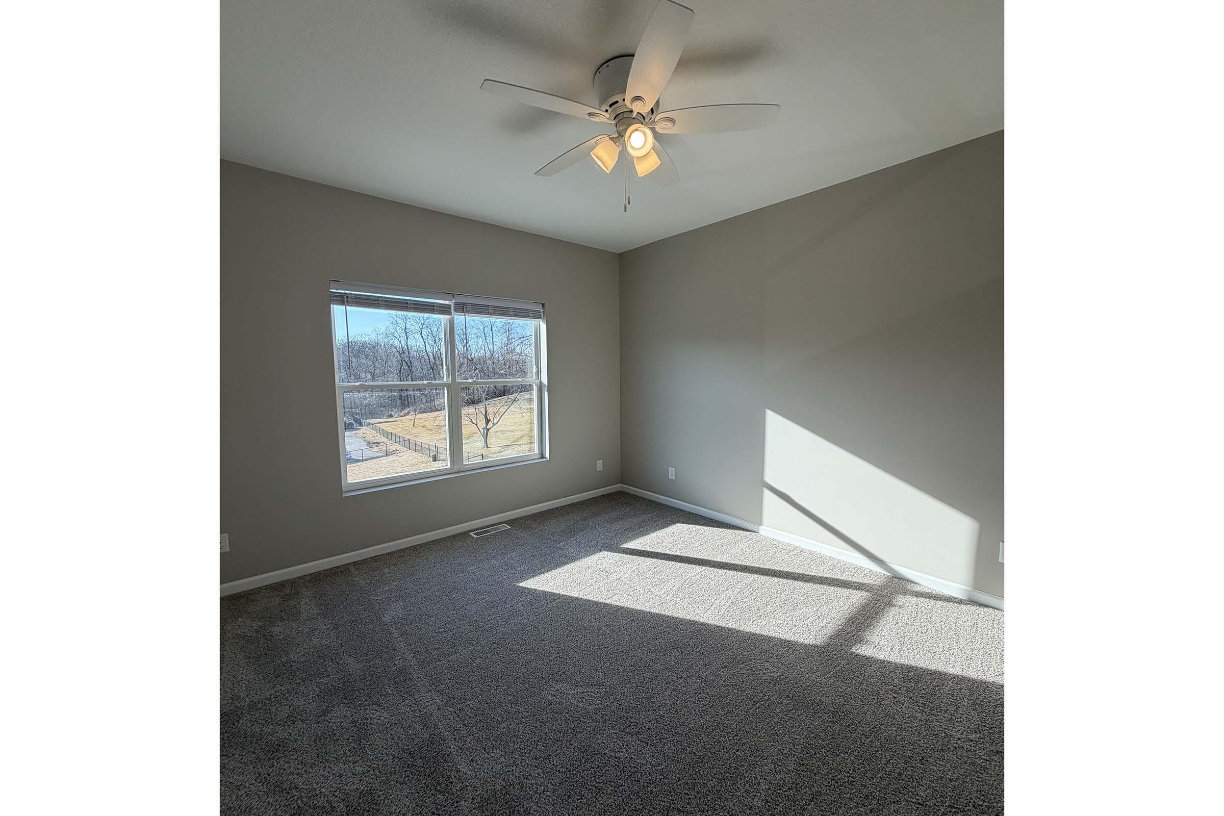 A well-lit room with light gray walls and carpet. It features a ceiling fan with lights, a large window allowing natural light, and a view of a grassy area outside. The room appears empty, creating a sense of spaciousness.