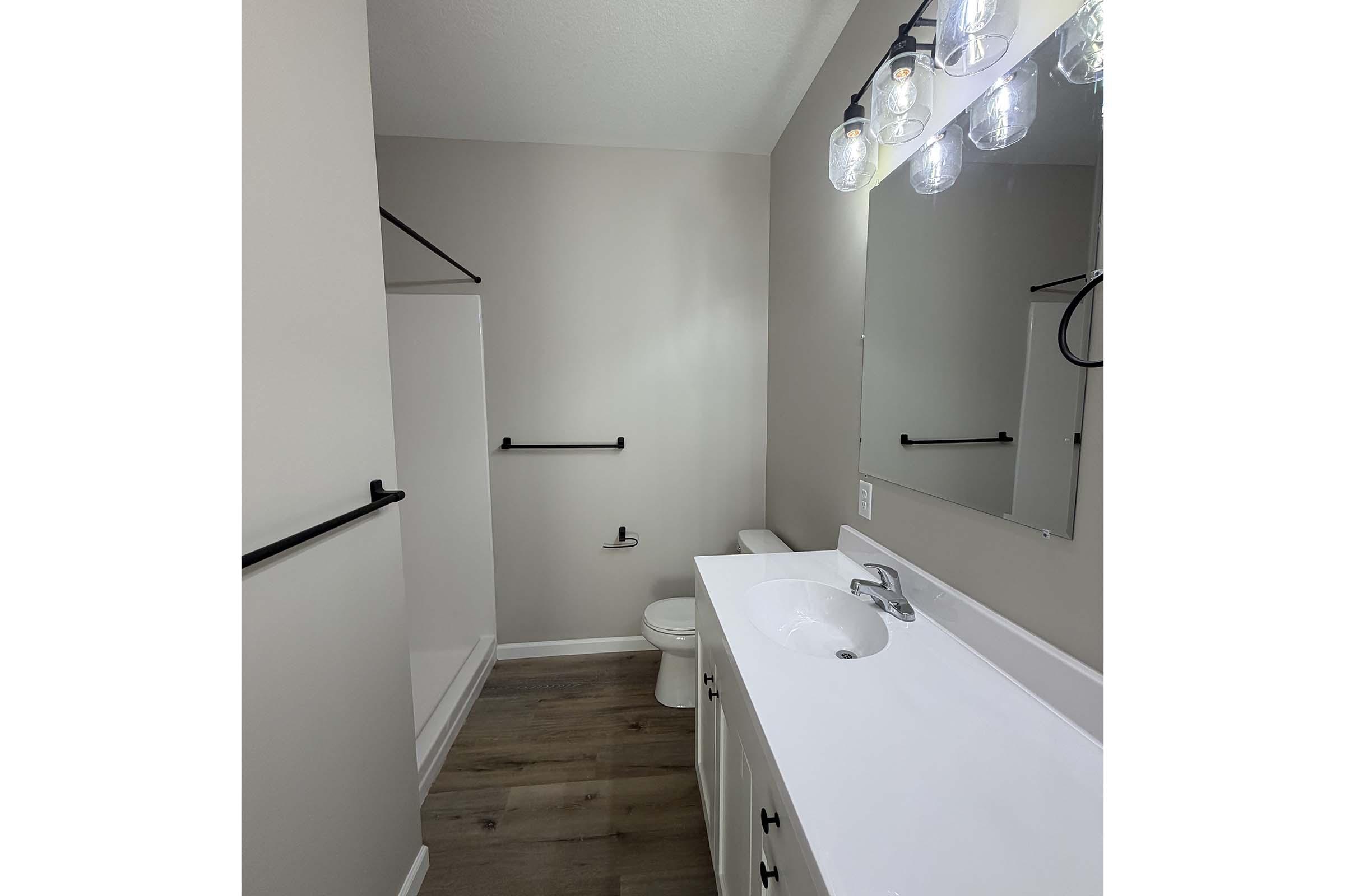 A modern bathroom featuring a white vanity with a sink, a large mirror above, and black hardware accents. There's a shower area with a glass door, a toilet, and wooden flooring, all in neutral tones. The lighting is provided by stylish hanging fixtures.