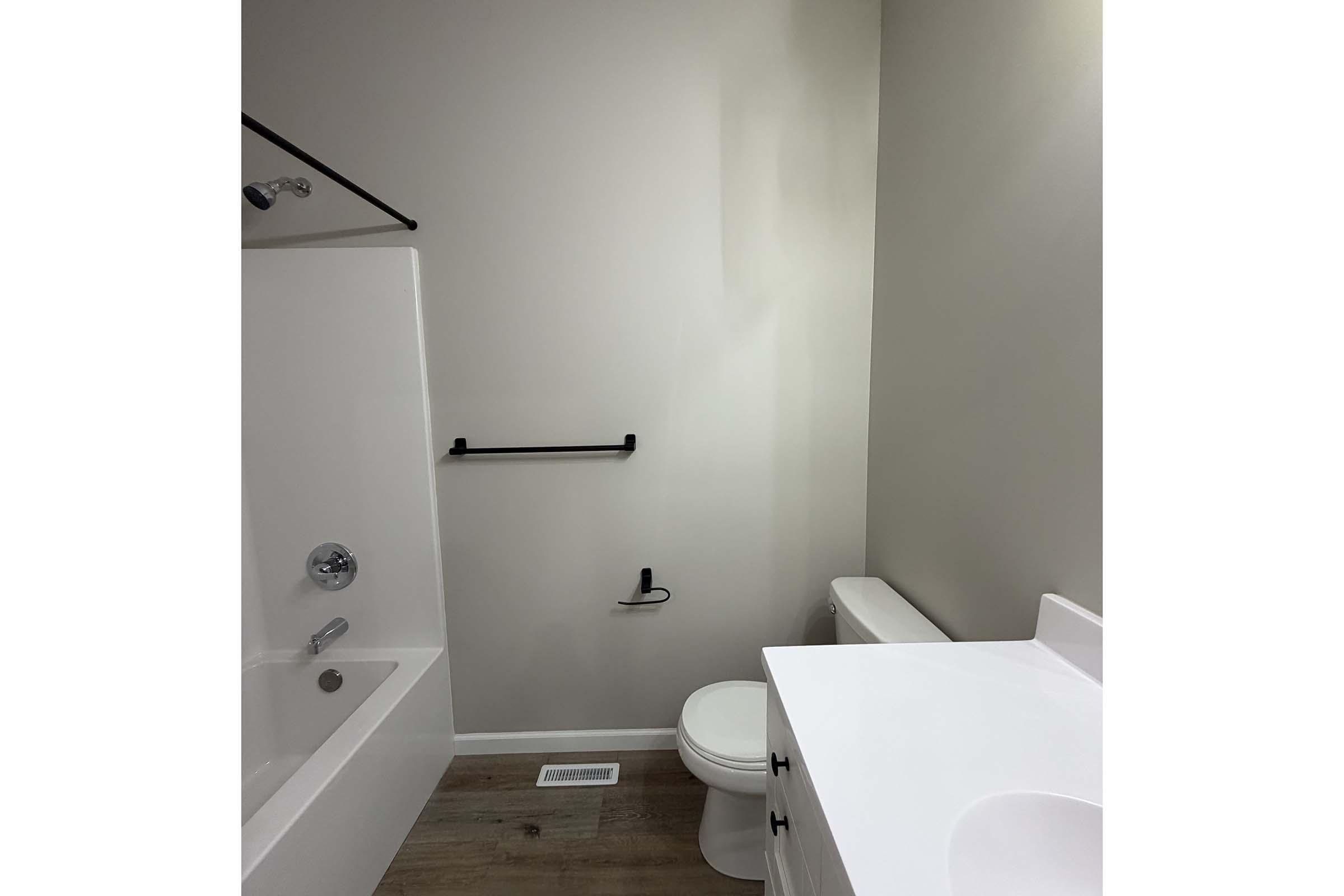 A clean and modern bathroom featuring a white bathtub with a showerhead, a black grab bar on the wall, a white toilet, and a sink with a white countertop. The walls are painted a light color, and the floor is made of wood-like material, creating a bright and spacious atmosphere.