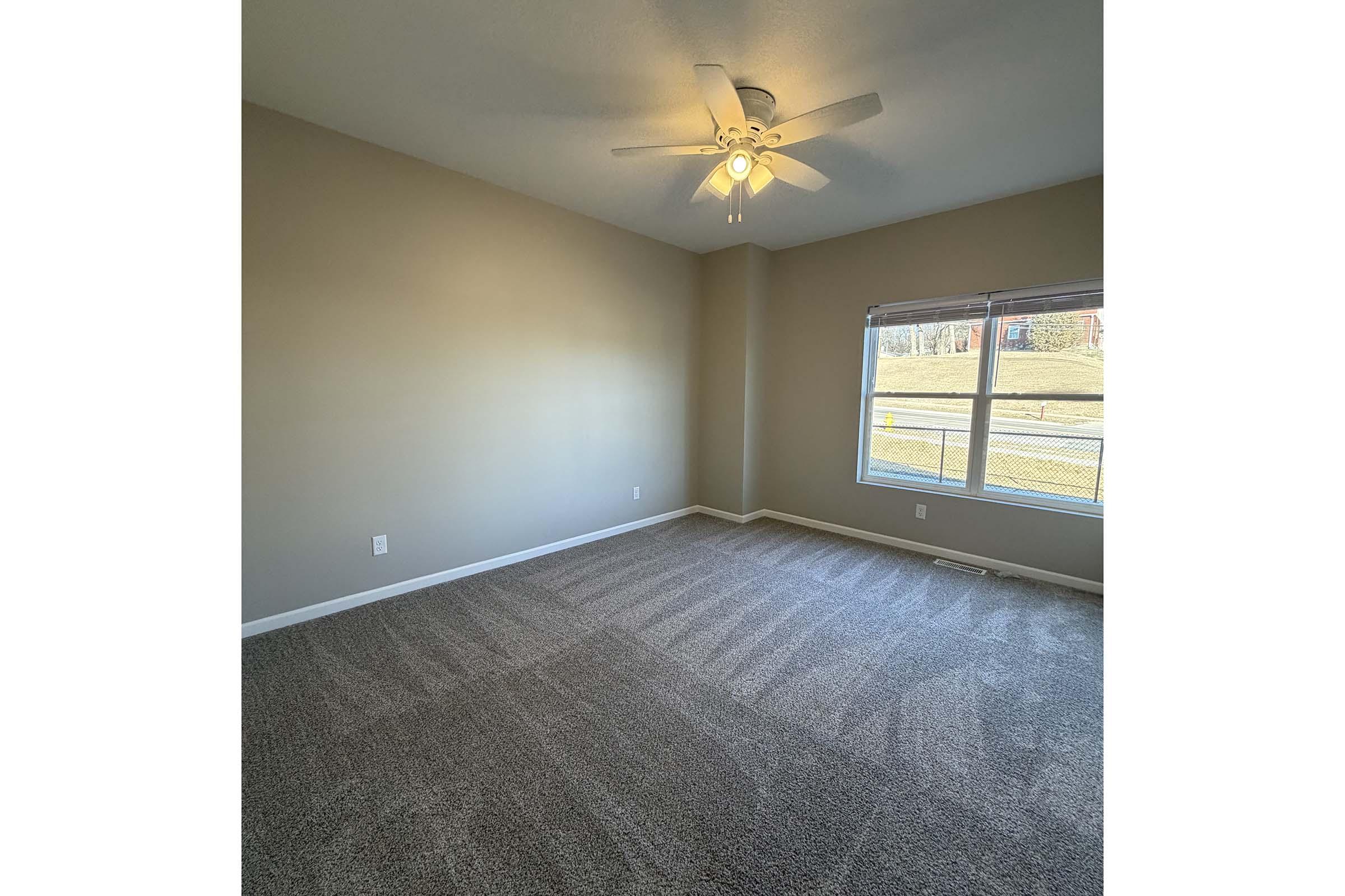 A spacious, empty room with beige walls and brown carpeting. A ceiling fan is installed, and there is a large window letting in natural light, offering a view of greenery outside. The room appears clean and well-maintained, suitable for various uses.