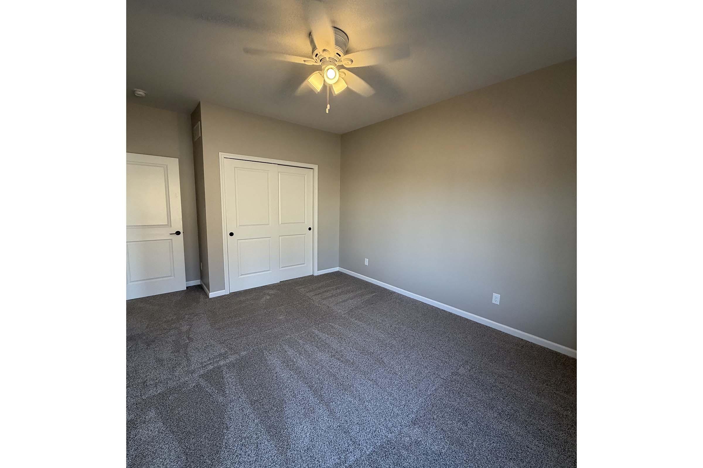 A spacious, empty room with light gray walls and plush carpet. A ceiling fan is visible, and there are two doors on the left, one leading to a closet. The room has natural light and a minimalist aesthetic, ideal for various uses such as a bedroom or office.