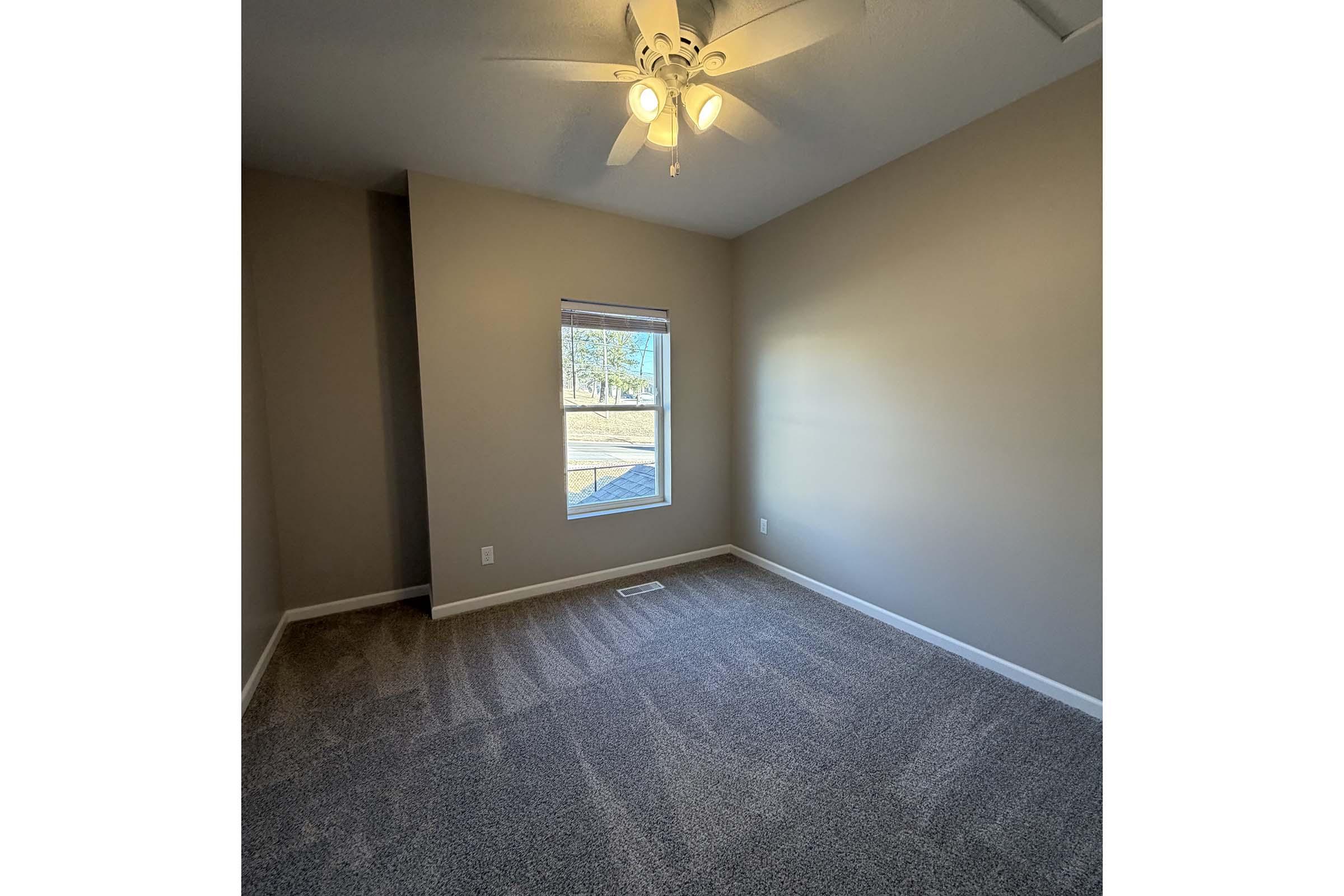 A well-lit, empty room with light brown walls and a ceiling fan. It features a window with a view of the outdoors and soft, plush carpeting on the floor. The room is spacious and uncluttered, inviting potential for various uses.
