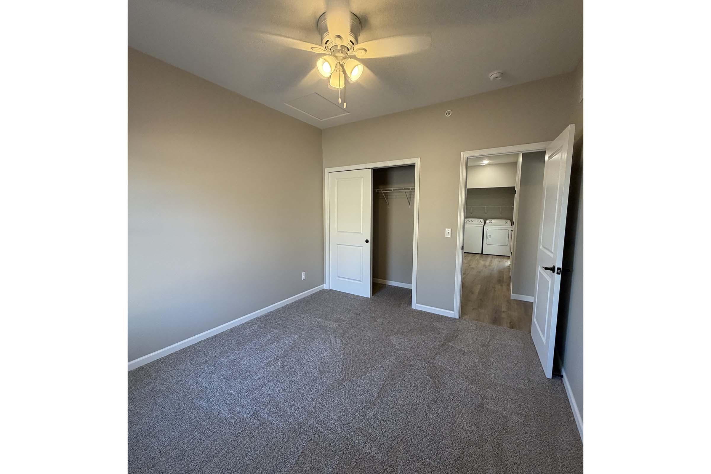 A well-lit room with neutral beige walls and carpet. It features a ceiling fan and two white doors: one leading to a closet and another that appears to open toward a laundry area. The room has a spacious feel, with natural light coming from an unseen window.