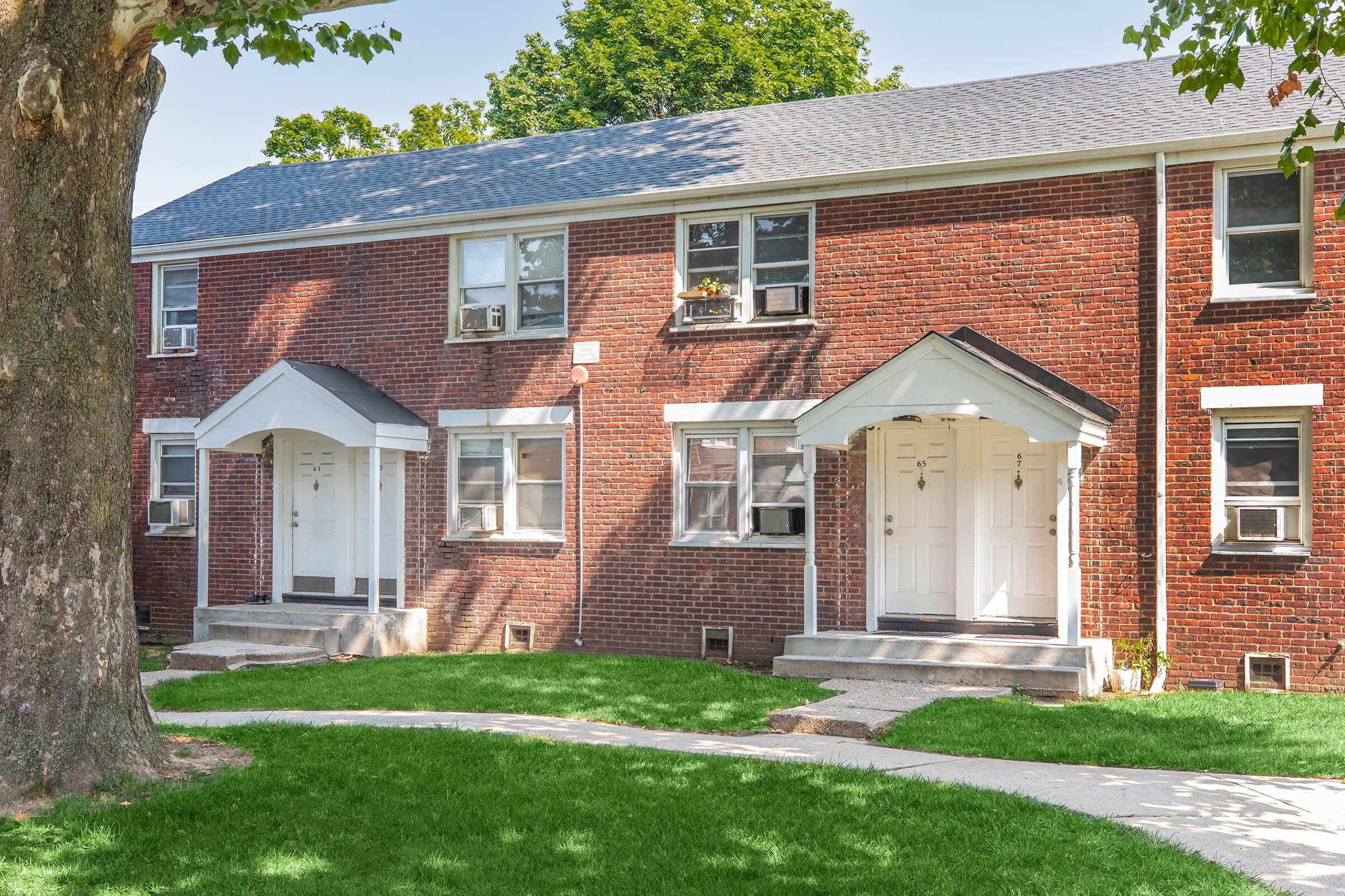 A view of a brick apartment building with two entrances, each featuring white doors and small front steps. The building is surrounded by green grass and a tree, with windows visible above the doors. The scene is well-lit, suggesting a sunny day.