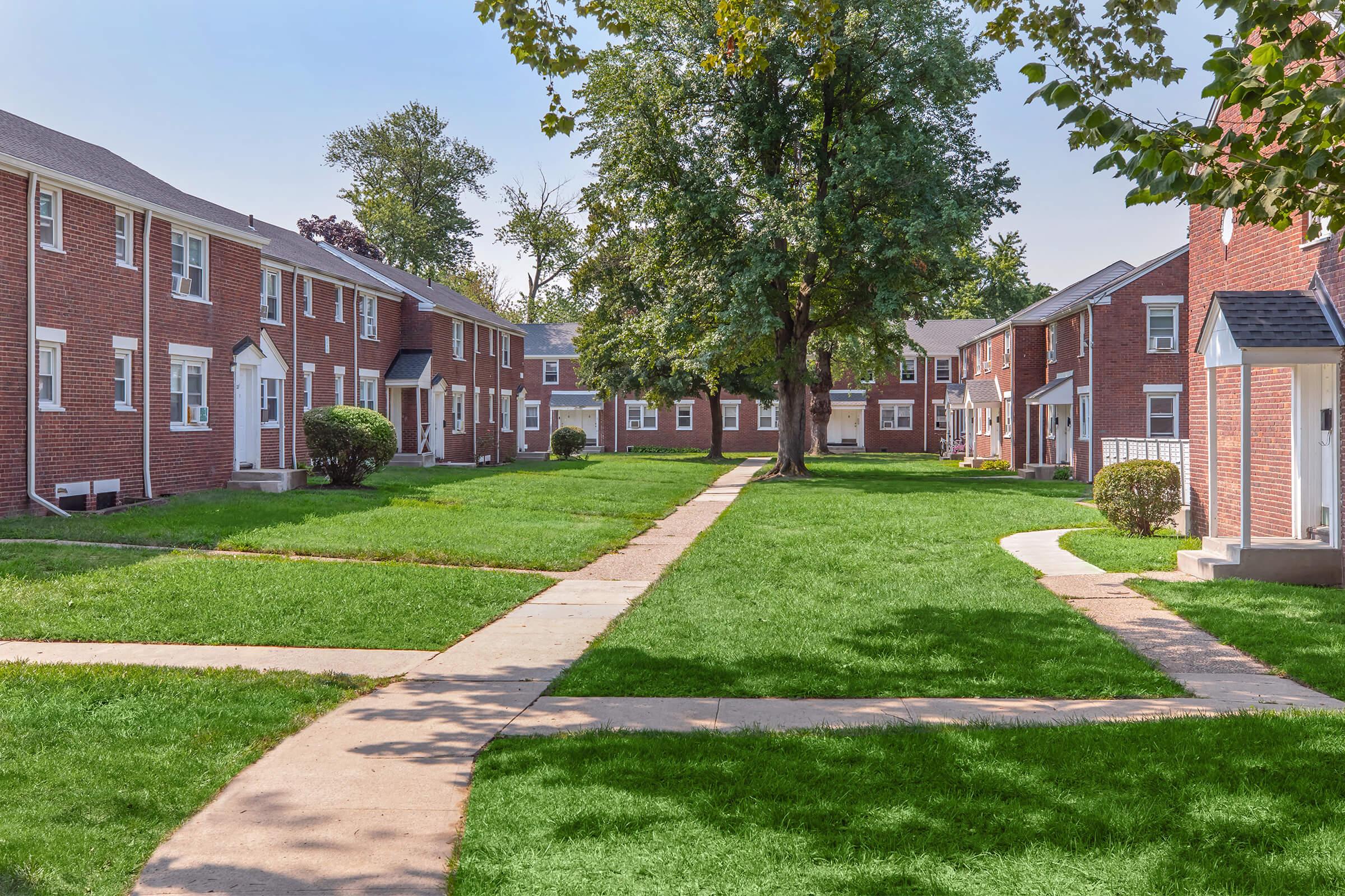 A view of a landscaped apartment complex featuring brick buildings arranged around a grassy courtyard. Pathways meander through the lawn, connecting the units, while trees provide shade in the background under a clear blue sky. The scene conveys a peaceful residential atmosphere.