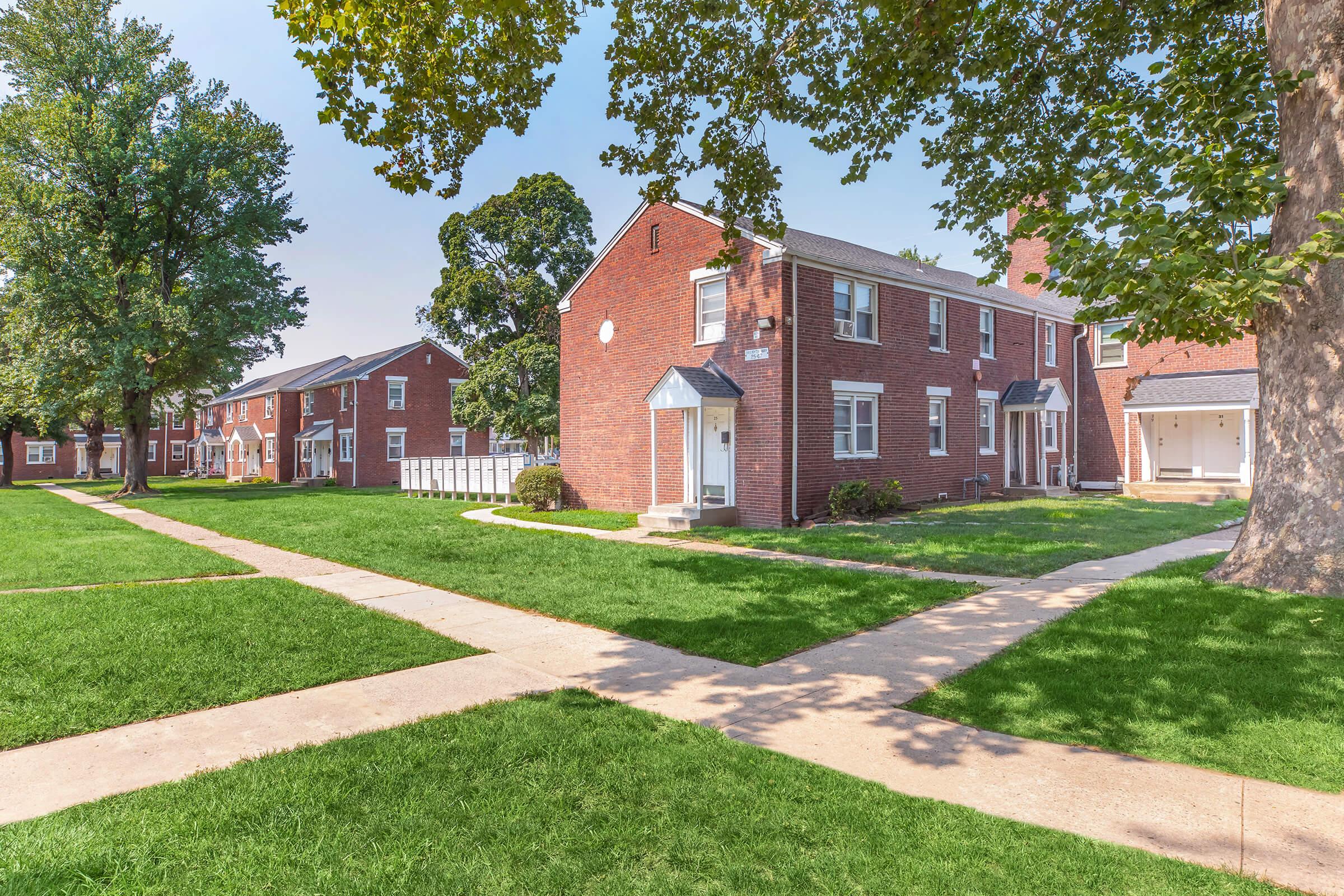 A well-maintained residential area featuring brick buildings with white trim. The layout includes grassy lawns and paved walkways, with trees providing shade. The scene captures a peaceful suburban atmosphere on a clear day.