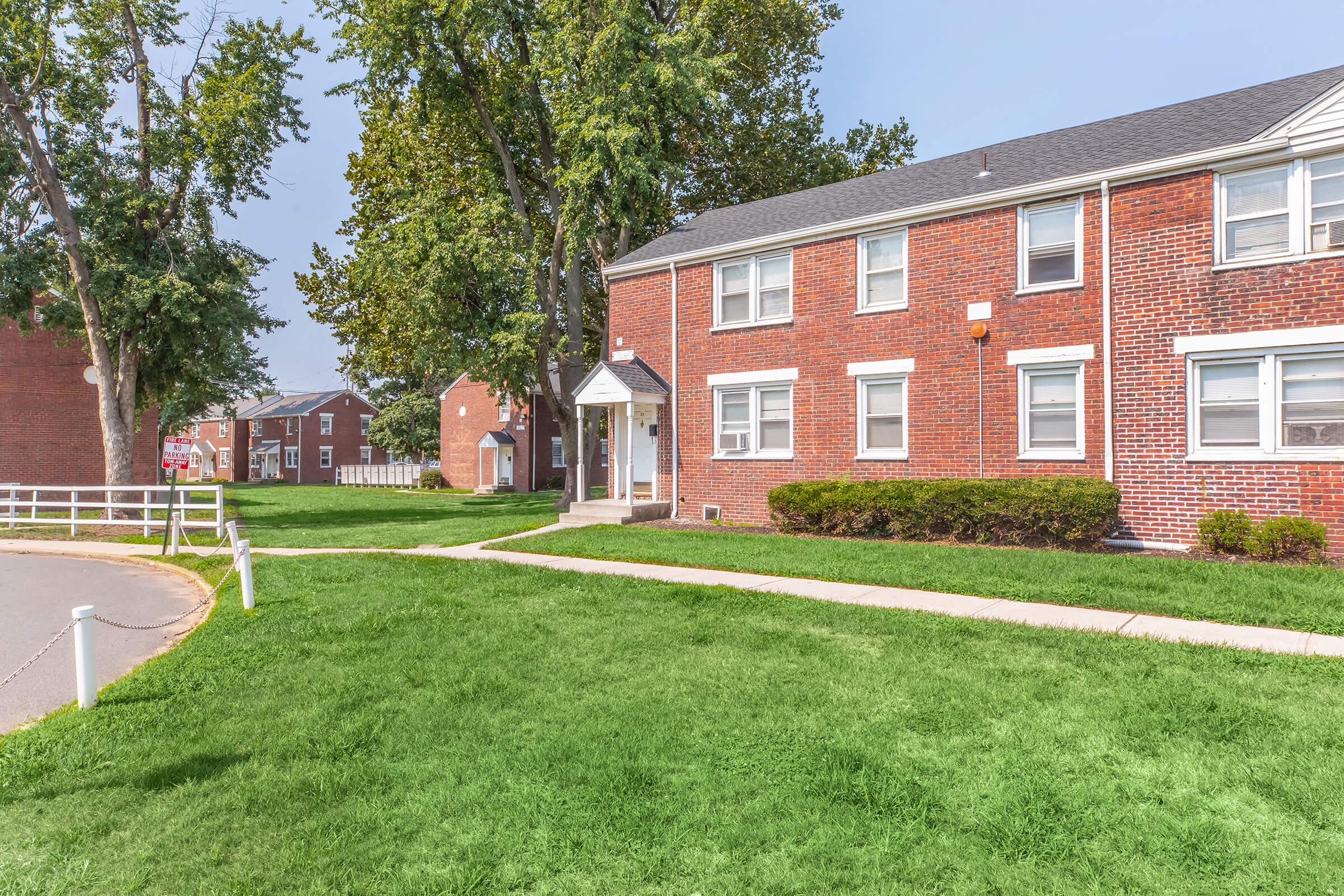 A brick apartment building with white trim located in a grassy area. Trees provide shade around the property, and there are additional buildings visible in the background. The pathway leads to the entrance, which features a small awning. The scene is bright and sunny with clear blue skies.