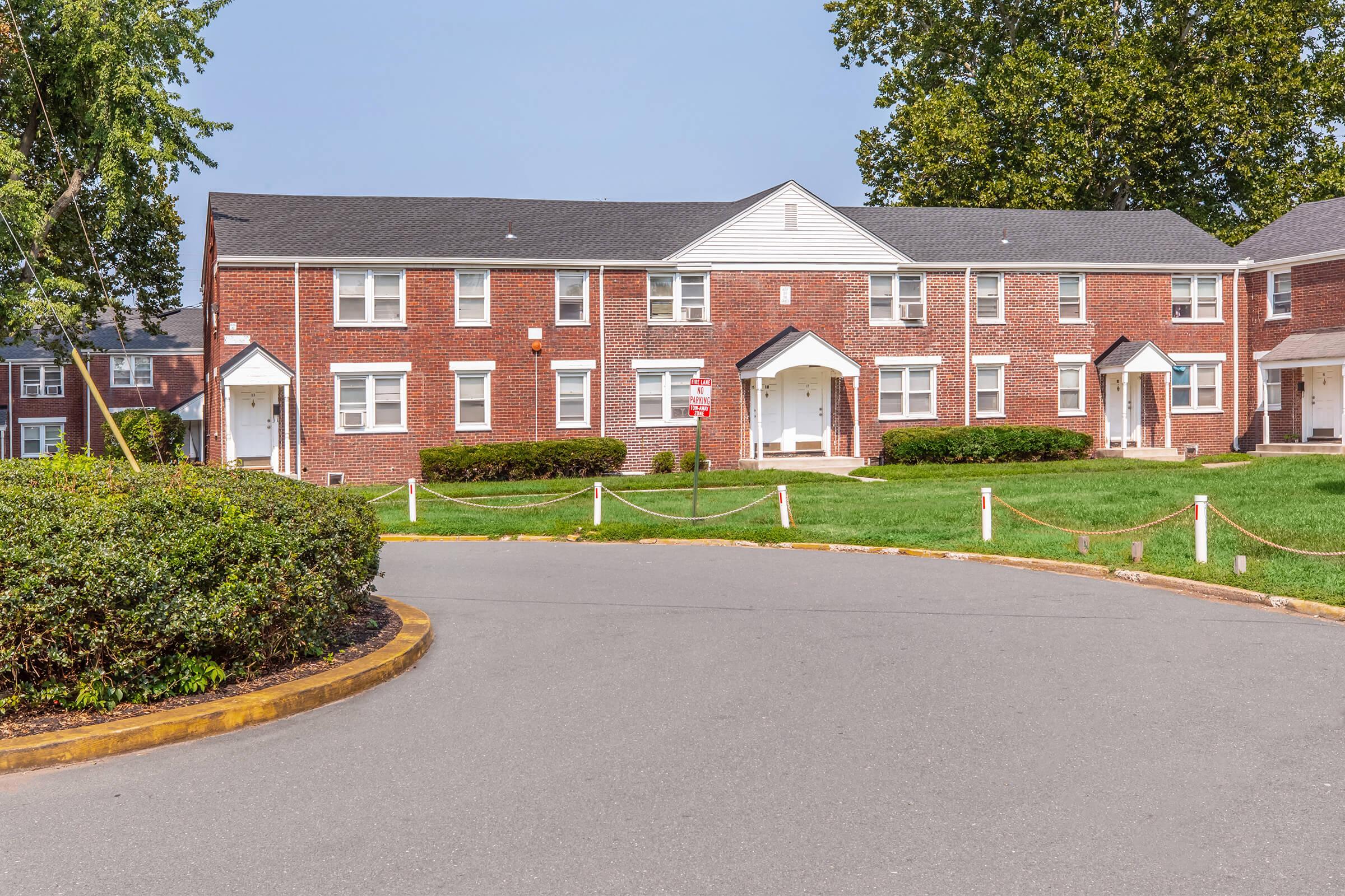 A brick apartment building featuring white trim, surrounded by well-manicured grass and bushes. The structure has multiple entrances with white doors and windows, under a clear blue sky. A paved circular driveway leads to the building, adding to the residential atmosphere.