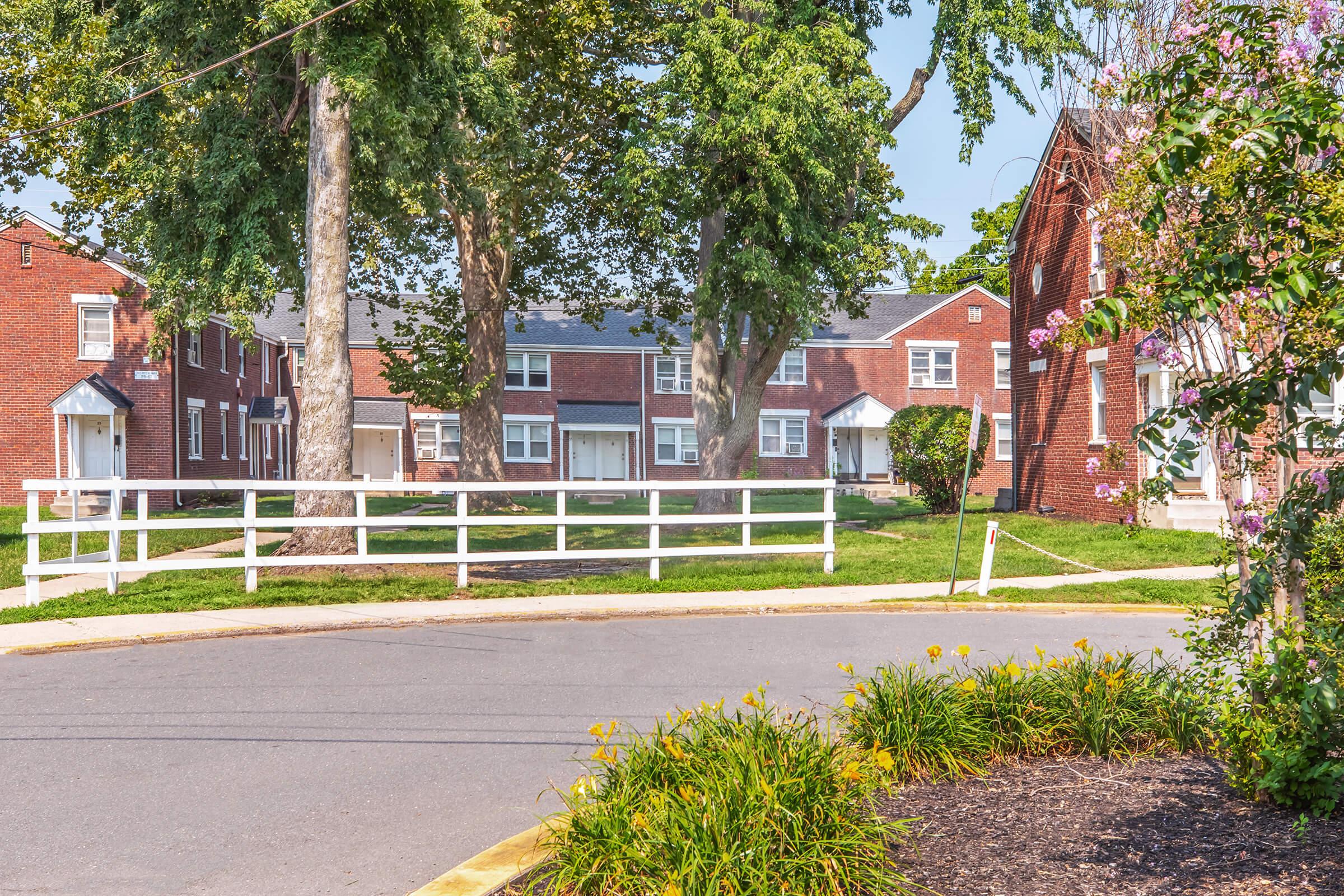 A peaceful residential area featuring several brick apartment buildings surrounded by lush green trees and well-maintained lawns. A white wooden fence runs along the pathway, with blooming flowers and a circular driveway in the foreground, creating a serene neighborhood atmosphere.