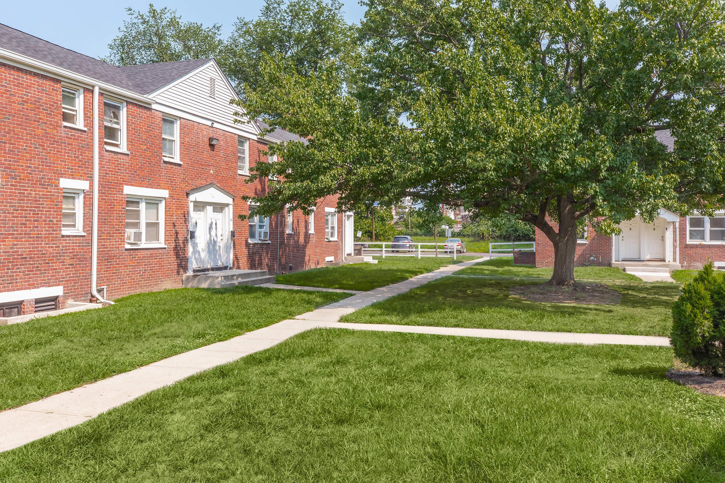 A view of a residential complex consisting of brick apartment buildings surrounded by green lawns and a large tree. There are concrete pathways leading to the entrances of the buildings, and the area appears well-maintained with sunlight illuminating the scene.