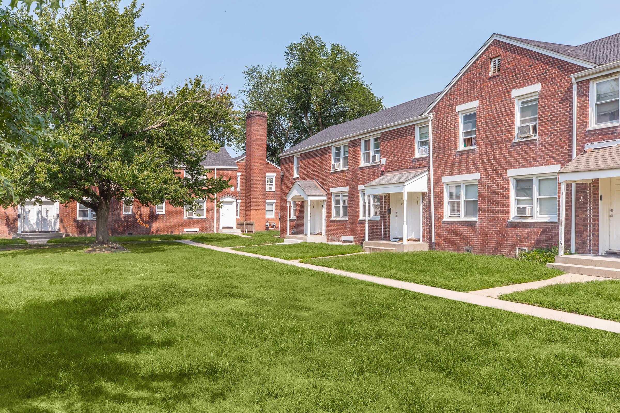 A residential area featuring two-story brick buildings with multiple doorways, surrounded by well-maintained green lawns and a few trees. The sky is clear and blue, indicating a sunny day. The layout suggests a community setting with a pathway connecting the buildings.