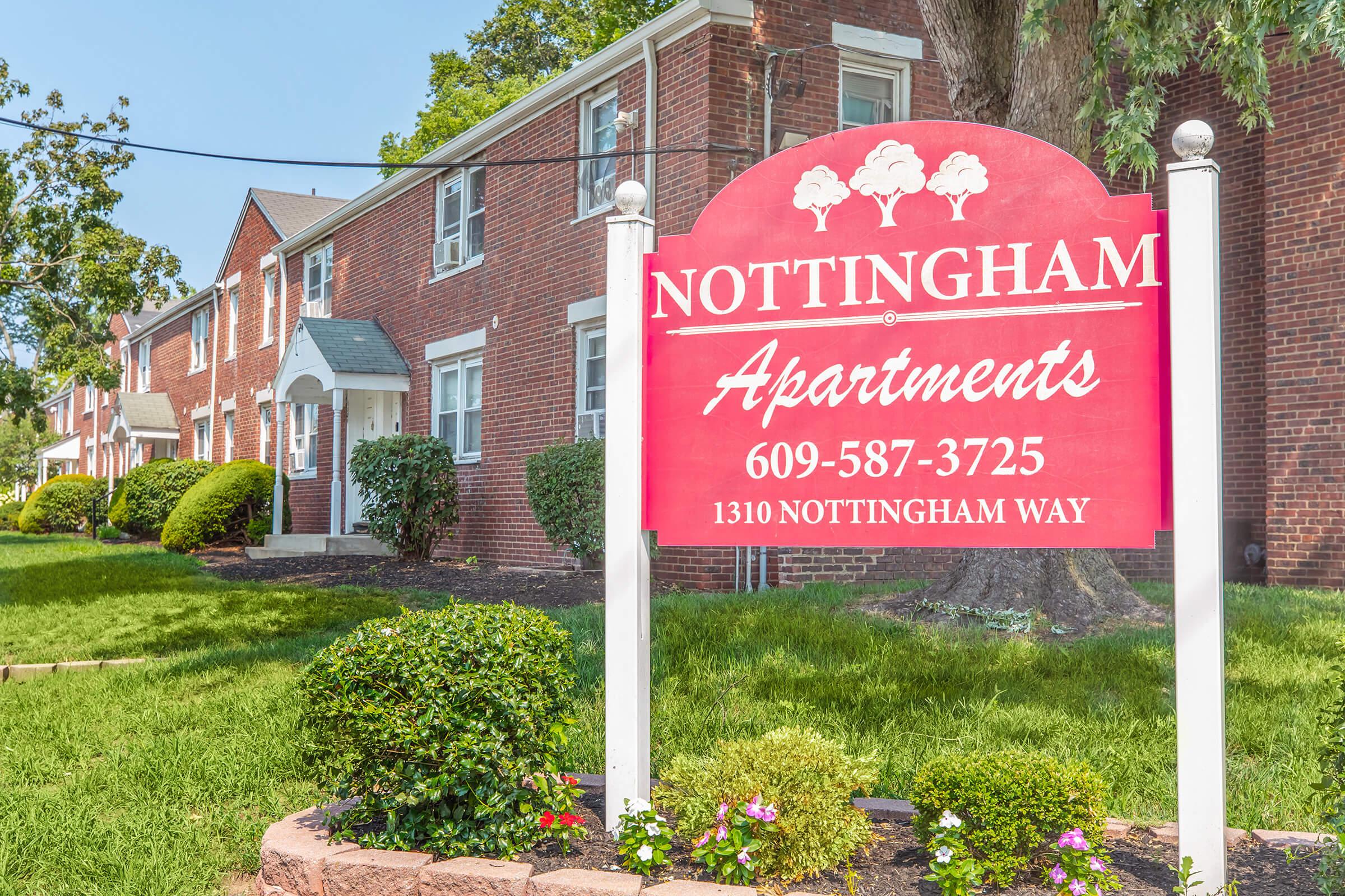 Sign for Nottingham Apartments, featuring a prominent red background with white text. The sign displays the phone number 609-587-3725 and the address 1310 Nottingham Way. The background includes a well-maintained pathway, green grass, and several trees and shrubs, indicating an inviting residential area.