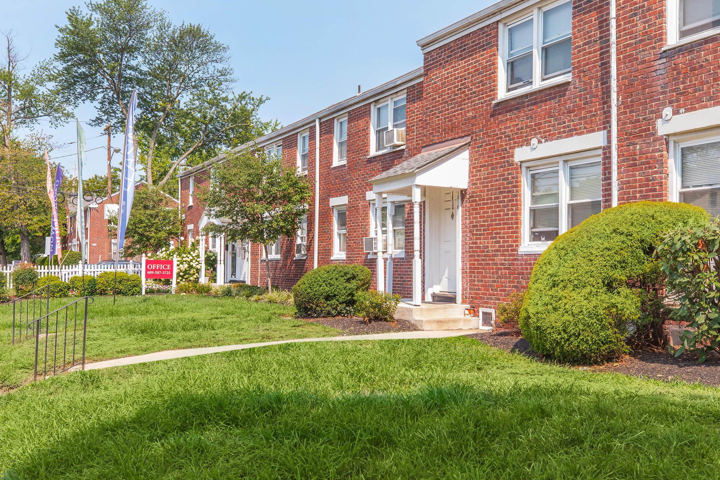 A view of a red brick residential building featuring several units, surrounded by well-maintained green lawns and shrubs. A sign labeled "OFFICE" is visible near the entrance. The scene is set under a clear blue sky, suggesting a warm, sunny day.