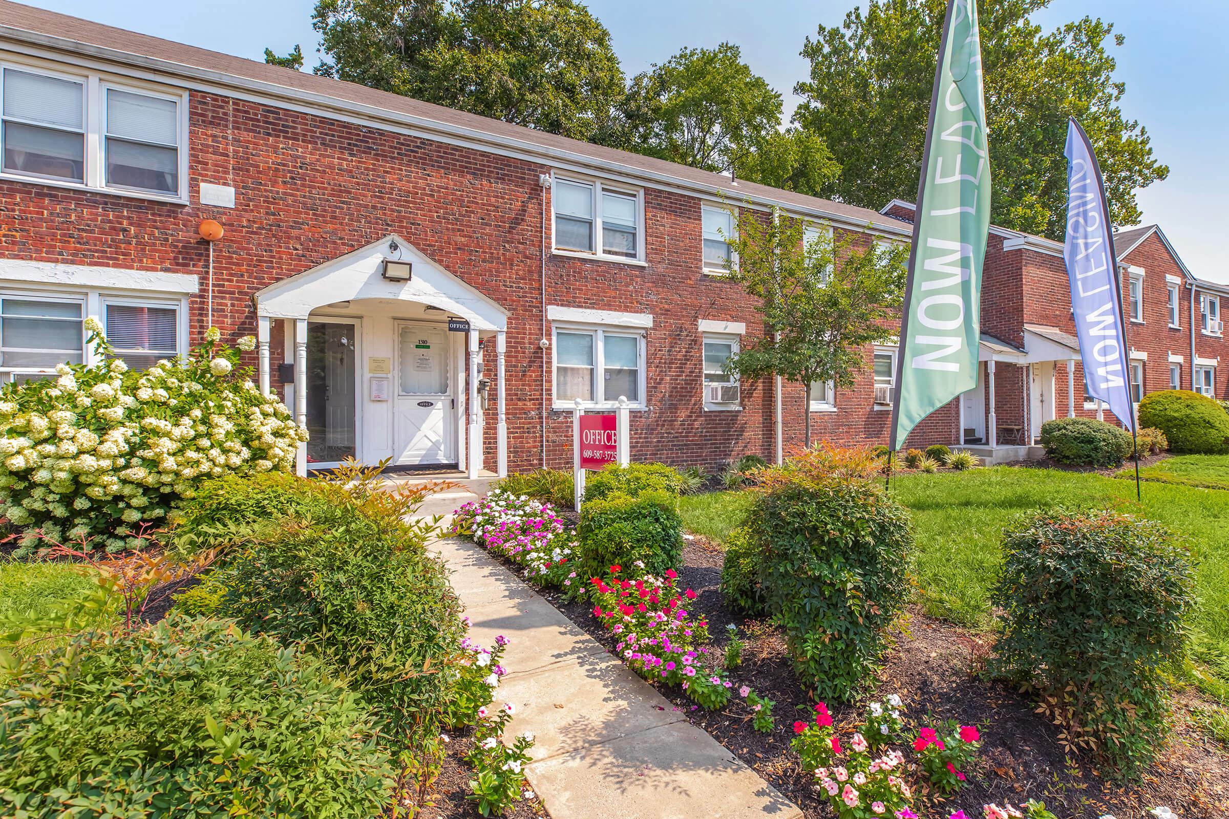 A brick apartment building with a sign reading "Now Leasing" in front. The entrance is flanked by colorful flags and surrounded by well-maintained flower beds and shrubs. A clear blue sky is visible in the background.
