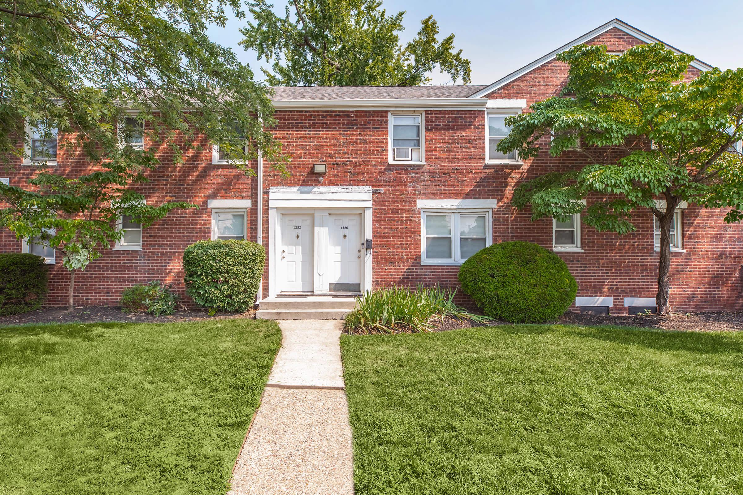 A brick apartment building featuring two front doors framed by greenery. A well-maintained lawn with shrubs lines the pathway leading to the entrance. The building has a simple, symmetric design with windows on either side of the doors, and trees provide shade in the vicinity.