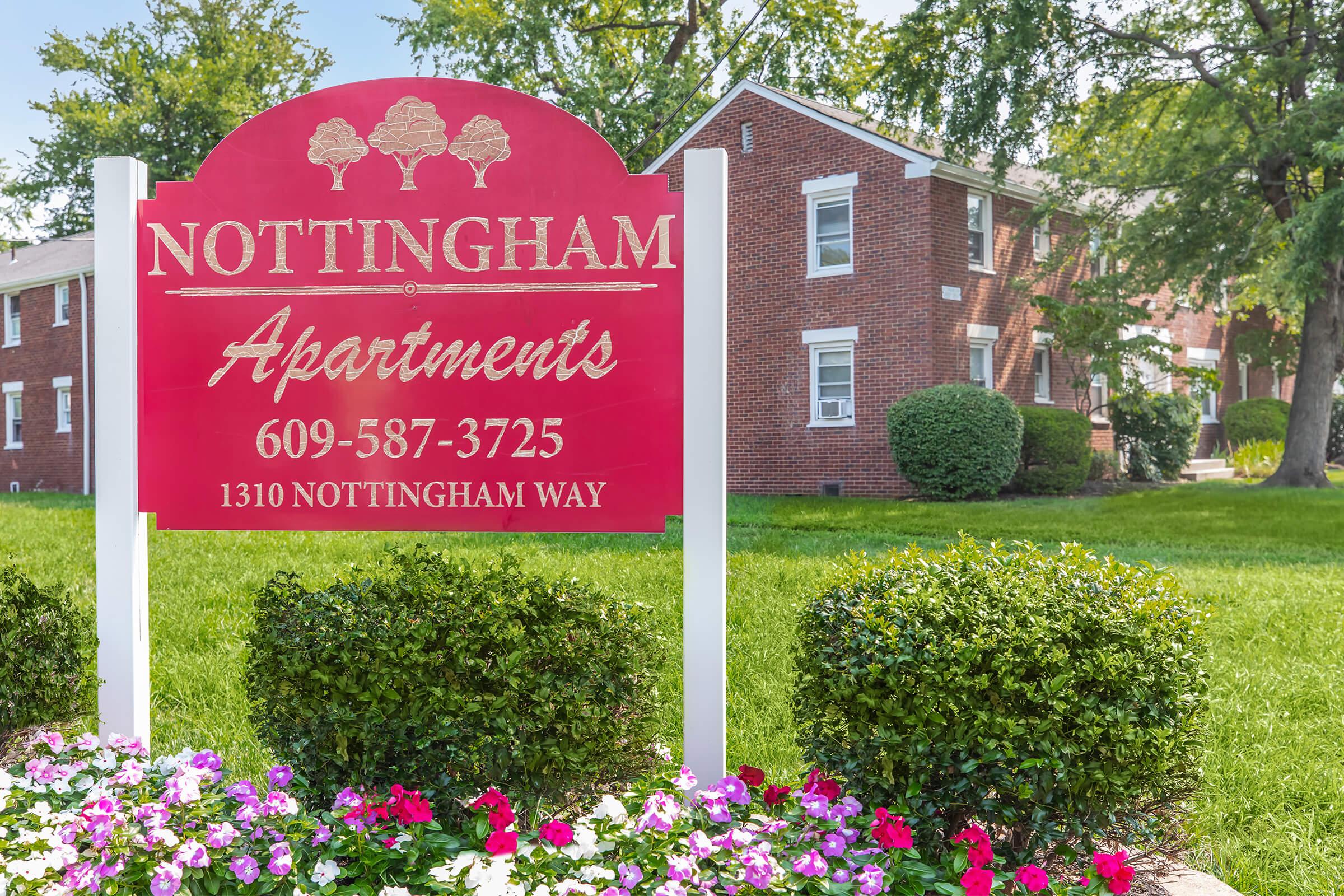 A red sign with white text reading "Nottingham Apartments" and the phone number "609-587-3725" is displayed in front of a brick apartment building. The surroundings include well-maintained green grass and flowering plants, creating a welcoming atmosphere.