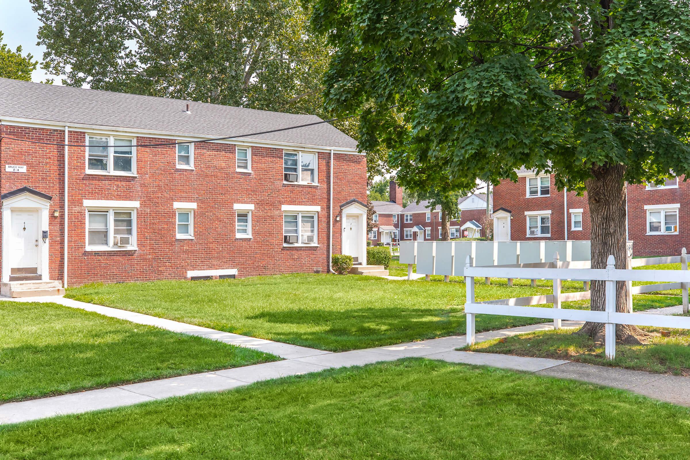 A view of a residential area featuring brick apartment buildings with white doors and windows. There is a well-maintained green lawn and a white picket fence surrounding the property. Trees provide shade, and the scene is bright and inviting, indicating a pleasant neighborhood environment.