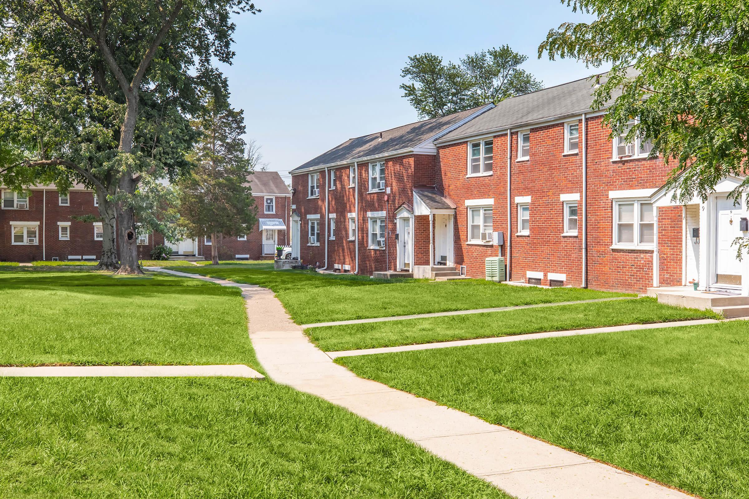 Brick apartment buildings arranged around a grassy courtyard with paved pathways leading to each unit. Several trees provide shade, creating a pleasant residential environment. The sky is clear, suggesting a sunny day.