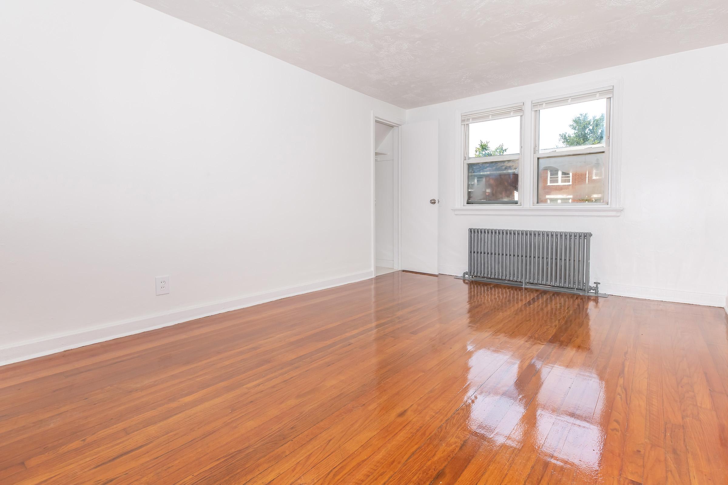 A bright, empty room with white walls and polished hardwood flooring. The room features a radiator near one wall, two windows allowing natural light, and a door leading to a closet. The space is clean and uncluttered, giving it a fresh and airy feel.
