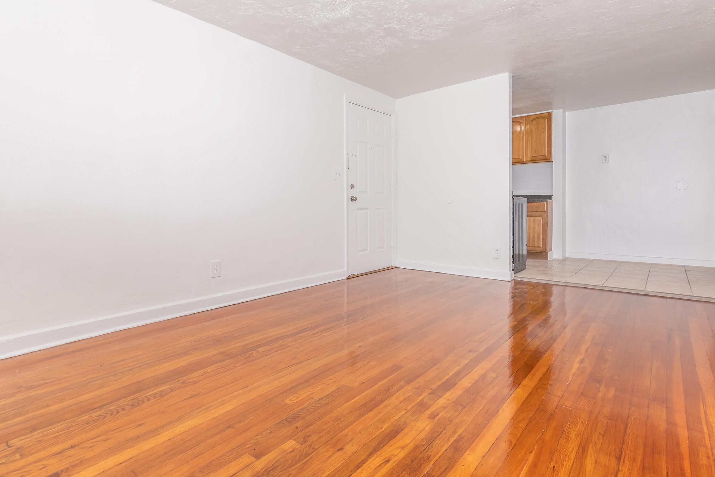 A bright, empty room with polished hardwood floors, white walls, and a closed door on the left. There is a doorway leading to a kitchen area in the background, which features light wood cabinets and tiled flooring. The space is well-lit, giving a fresh and clean appearance.