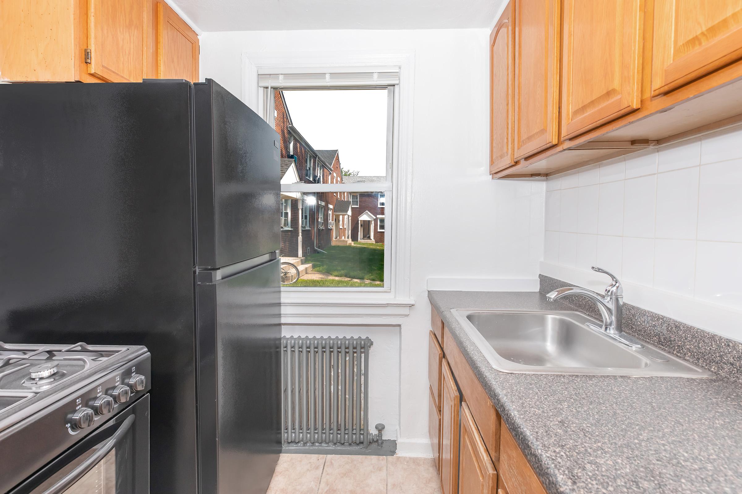 A small kitchen featuring a black refrigerator, a silver sink with a single faucet, and wooden cabinets. There is a window providing a view of a green outdoor area. A gas stove is visible on the left, and a radiator is located near the sink. The countertops have a dark stone appearance.