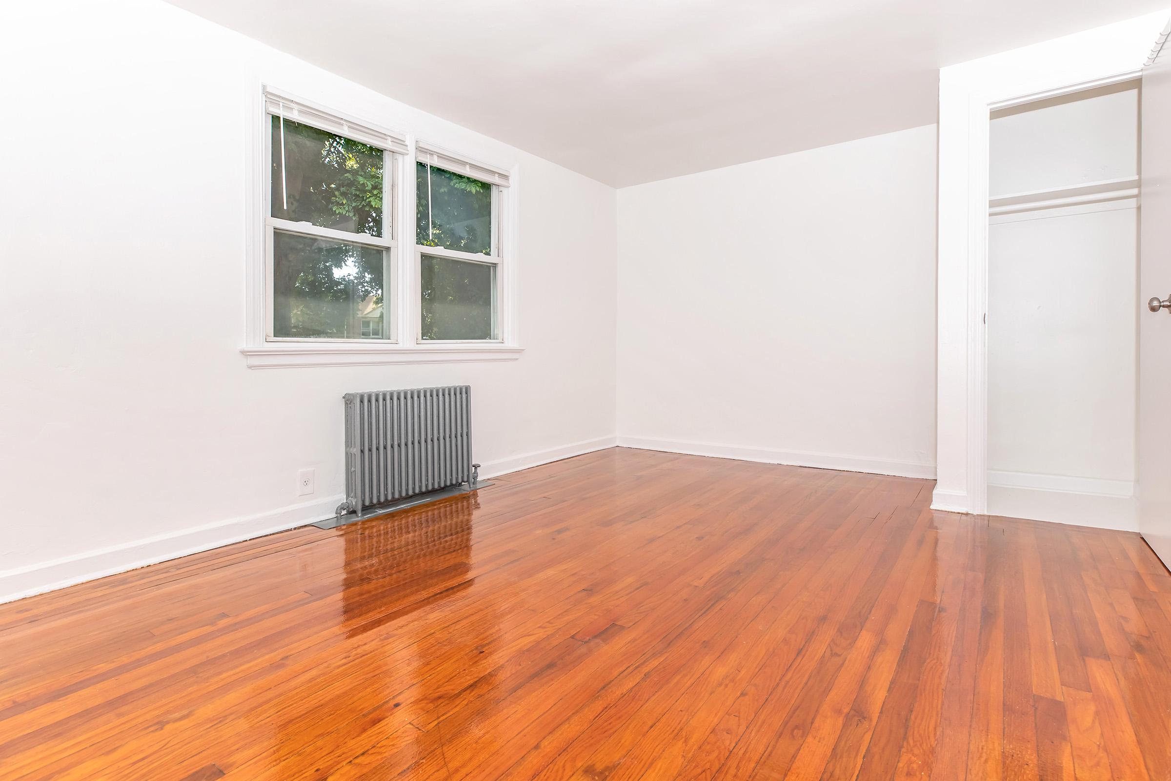A bright, empty room with hardwood flooring, featuring two windows letting in natural light and a wall-mounted radiator. One corner shows a closet with a door slightly ajar. The walls are painted white, creating a clean and spacious appearance. Ideal for personalizing or decorating.