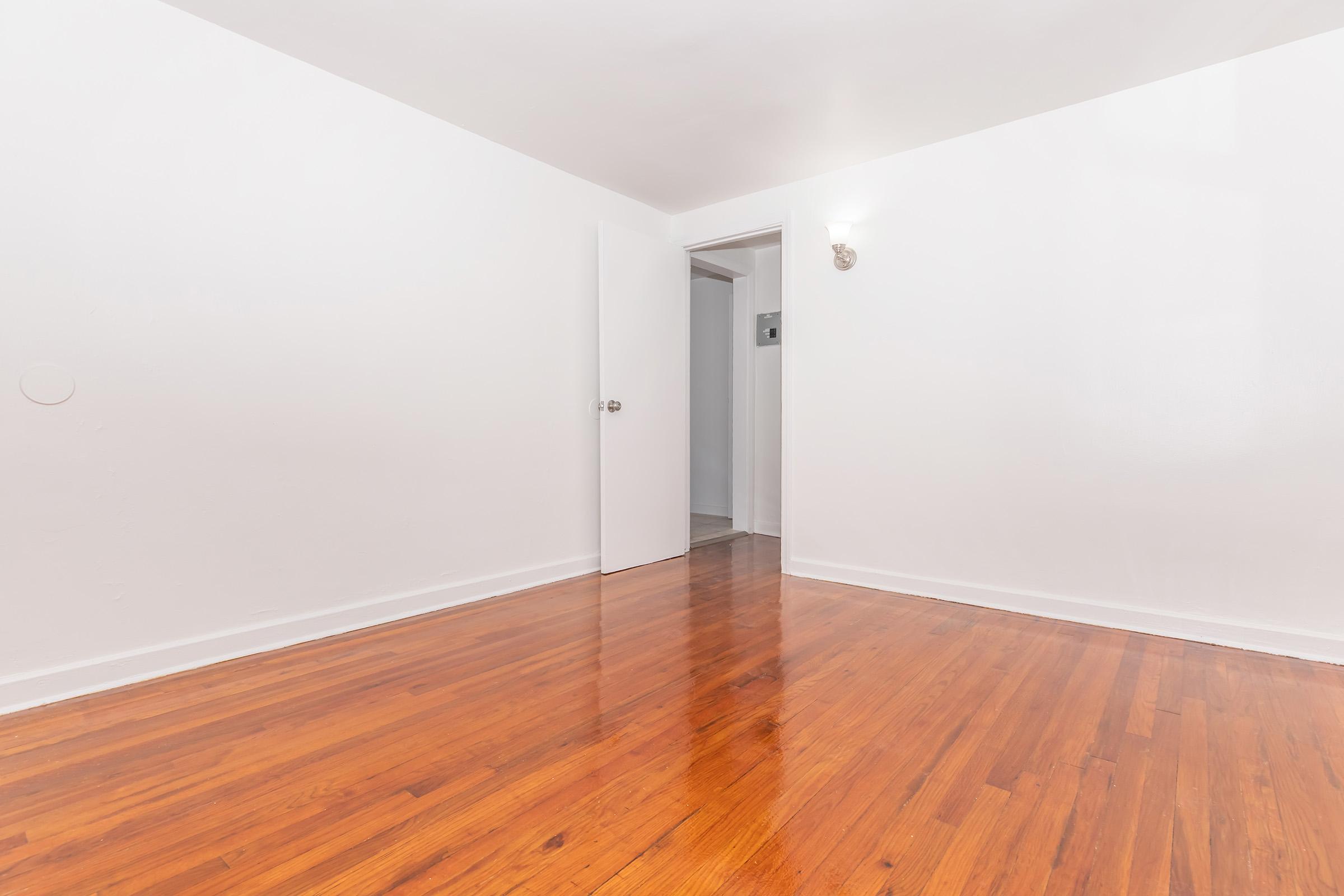 A bright, empty room with white walls and polished hardwood flooring. A door is slightly ajar, leading to another space. Soft lighting fixtures on the wall provide illumination, creating a clean and spacious atmosphere. The room is devoid of furniture, emphasizing its openness.