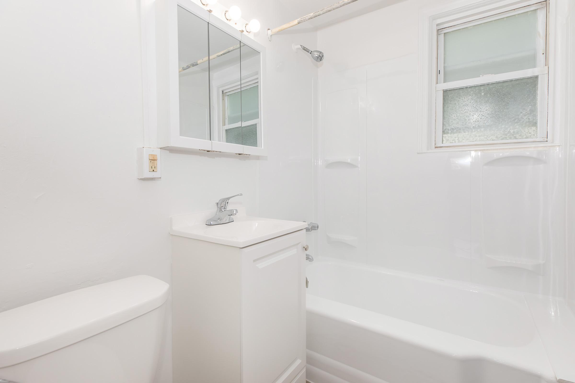 A clean, modern bathroom featuring a white bathtub, a compact sink unit, and a toilet. A wall-mounted mirror with lights above is present, along with a window allowing natural light. The walls and fixtures are bright and minimalistic, creating a spacious feel.