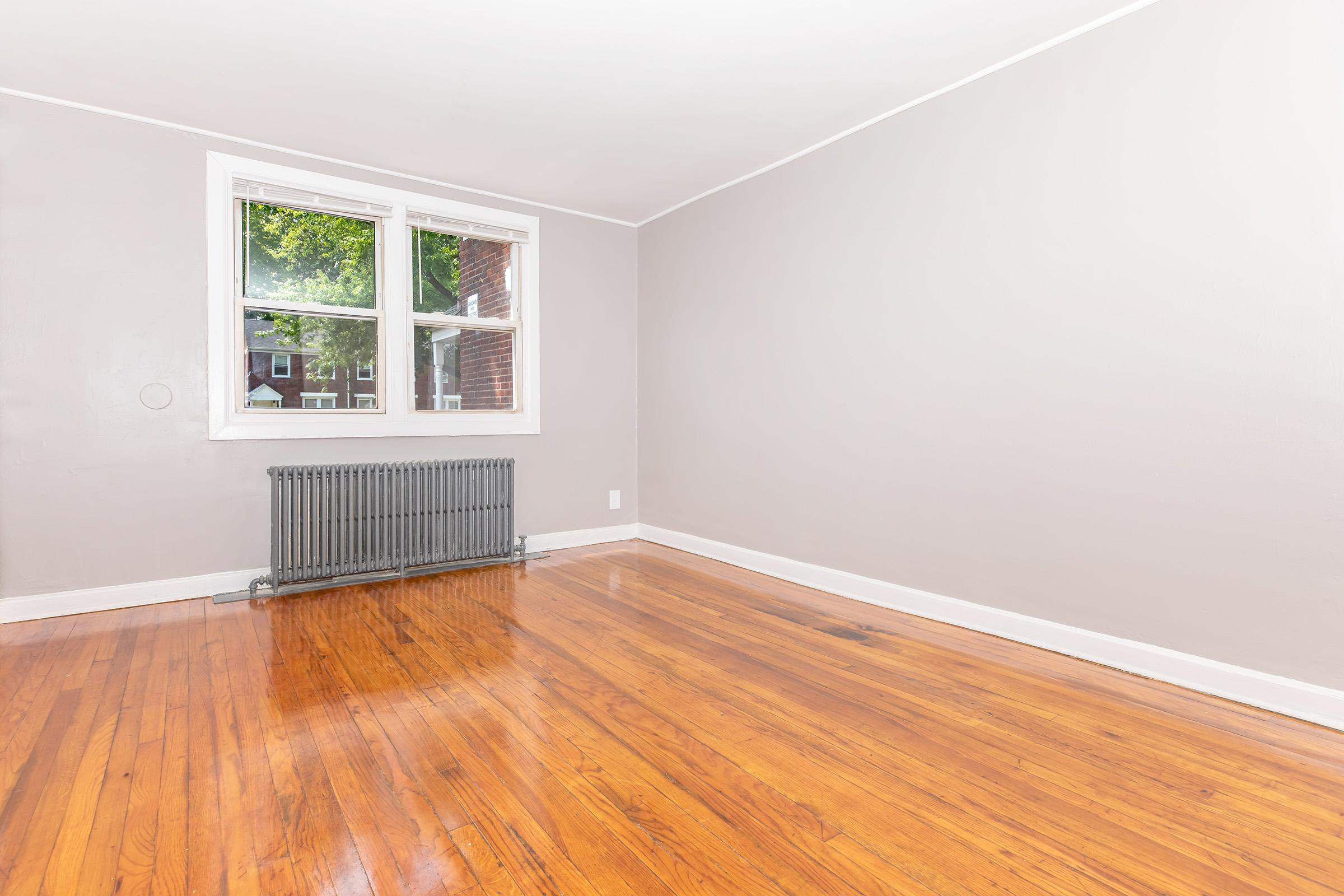 A vacant room with light gray walls and polished hardwood floors. A single window lets in natural light, reflecting off the wooden surface. A vintage radiator is mounted beneath the window, adding to the room's charm. The space appears clean and inviting, ready for decoration.