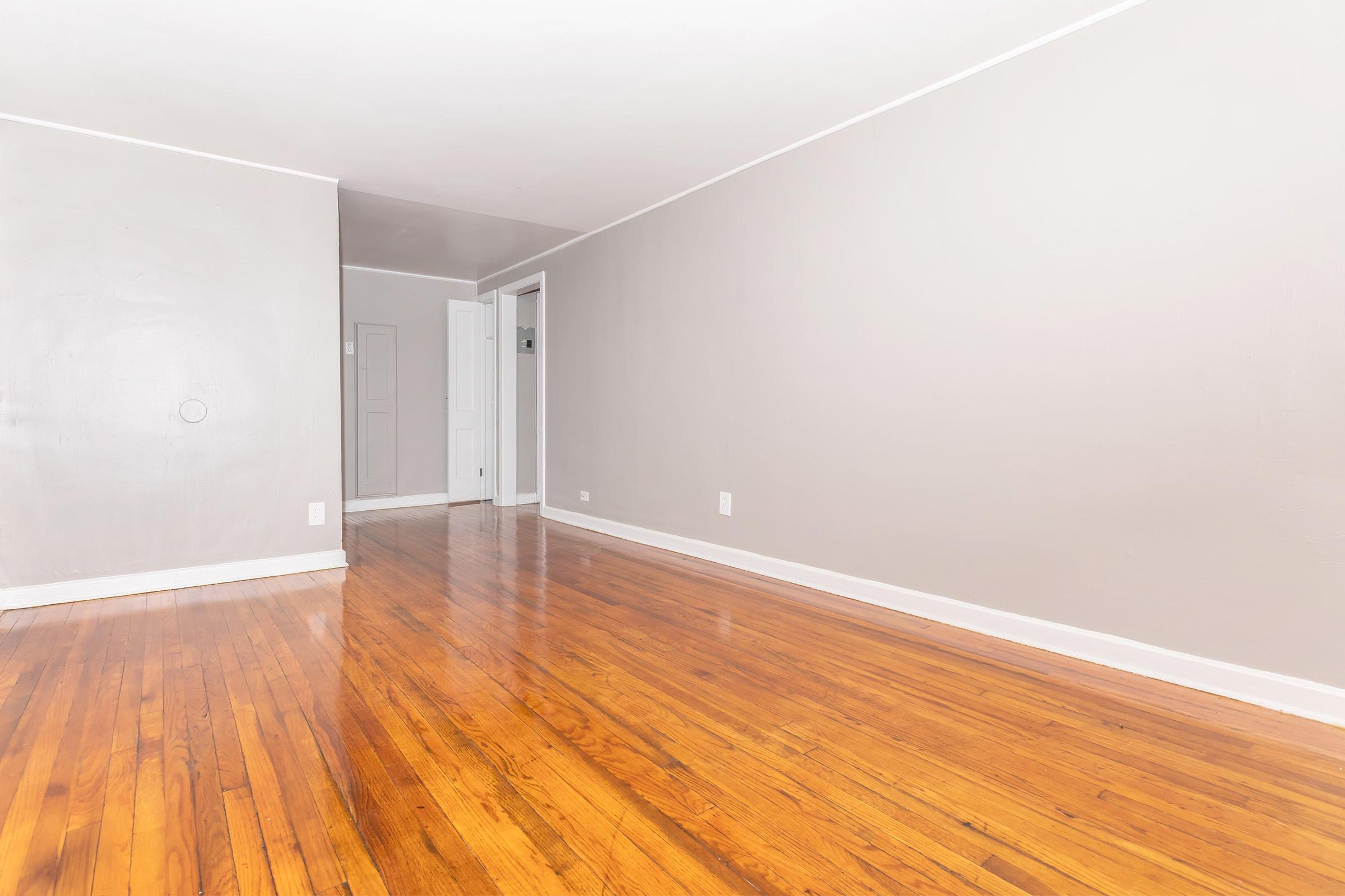 Spacious interior of a room with hardwood floors, light gray walls, and minimal decoration. A door can be seen on the right leading to another area, while the left side features a circular object on the wall. The overall atmosphere is bright and uncluttered, suggesting a clean and open living space.