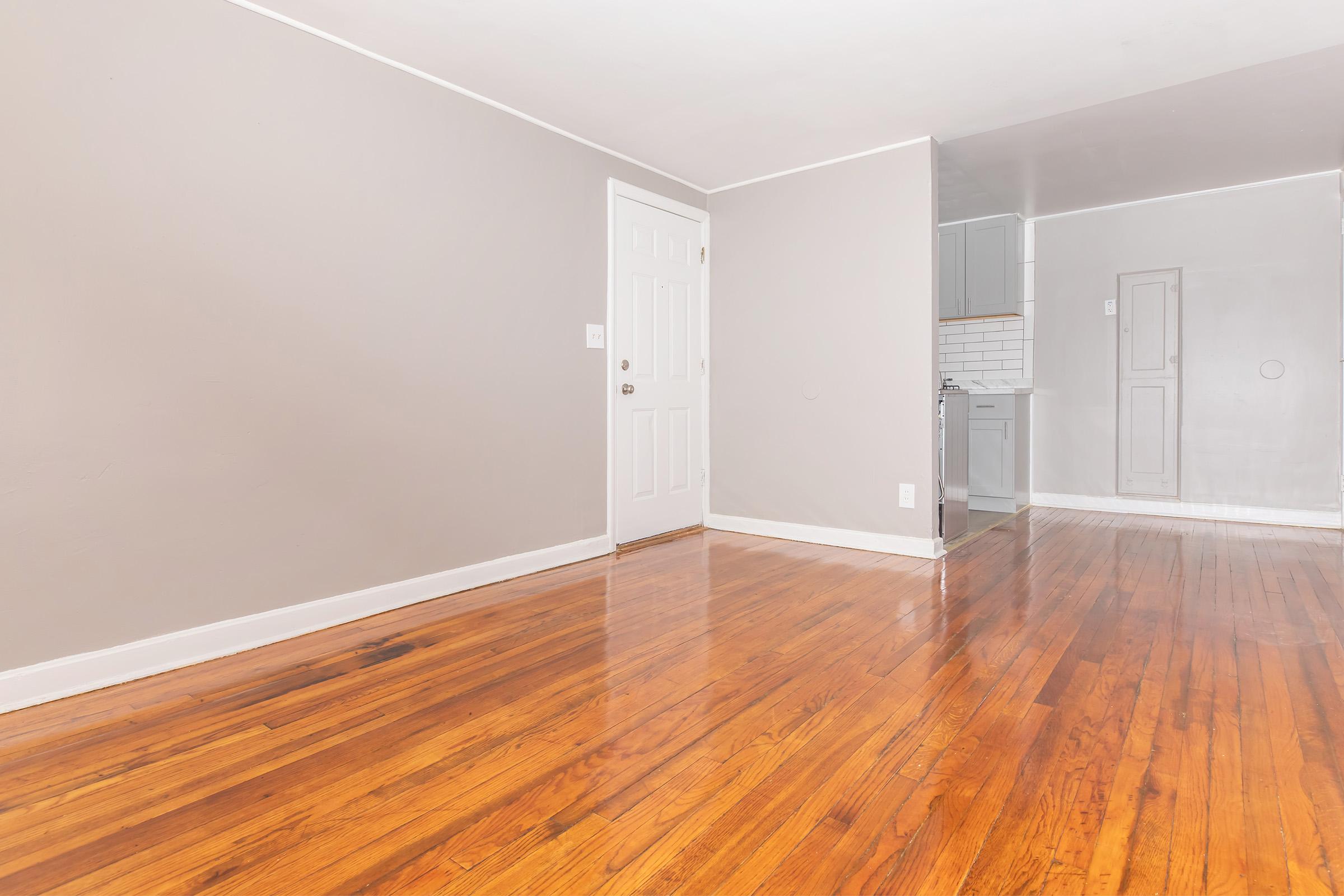 A spacious, empty room with polished hardwood floors and light gray walls. A white door is visible on the left, leading to an exterior entrance. The right side of the image shows a small kitchenette area with light cabinetry and a tiled backsplash, creating a clean and modern appearance.