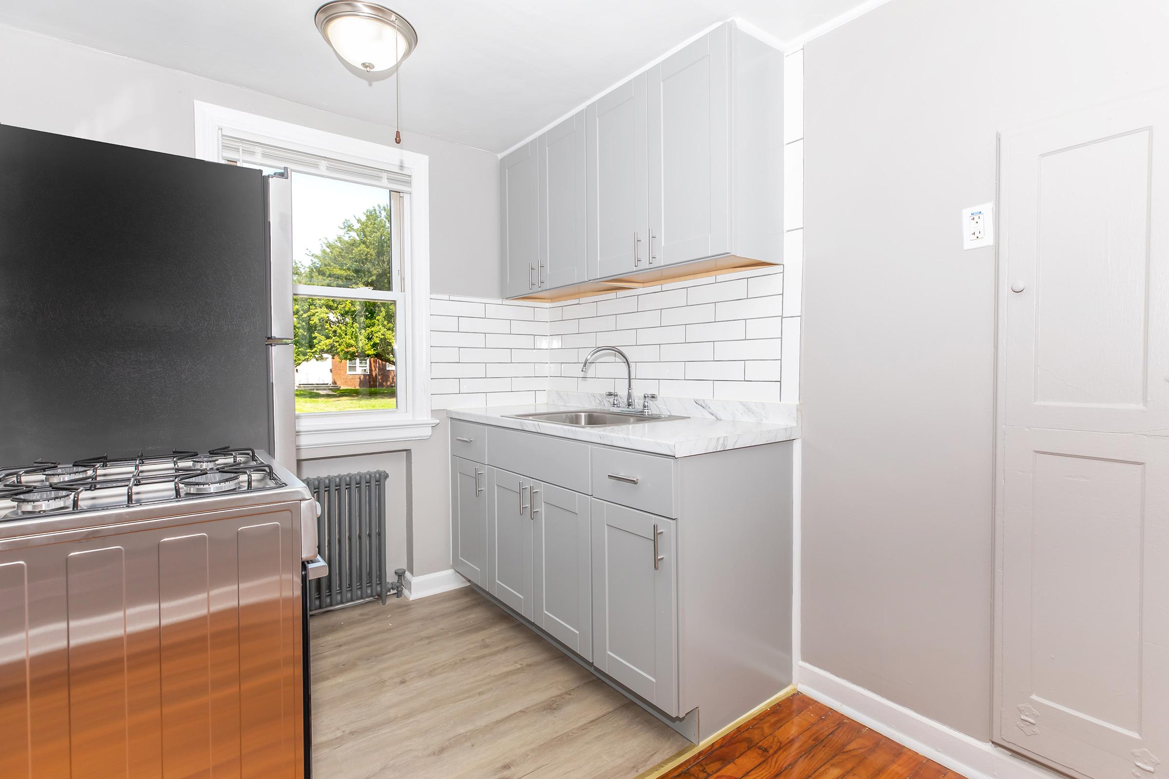 Modern kitchen featuring gray cabinets, a white tiled backsplash, and a marble countertop. Includes a stainless steel stove and refrigerator, a window providing natural light, and a wooden floor. A radiator is visible near the wall.