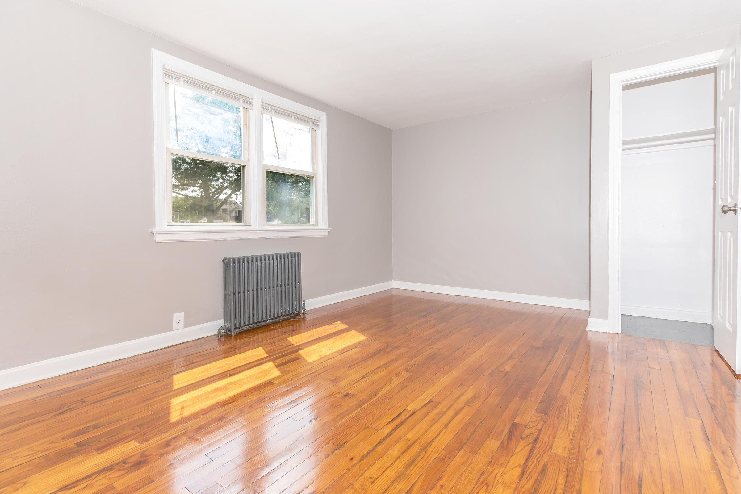 A clean, empty room with light gray walls, hardwood flooring, and two windows allowing natural light. A small radiator is positioned beneath the windows, and there’s an open closet visible on the right side of the image. The room feels spacious and well-lit.