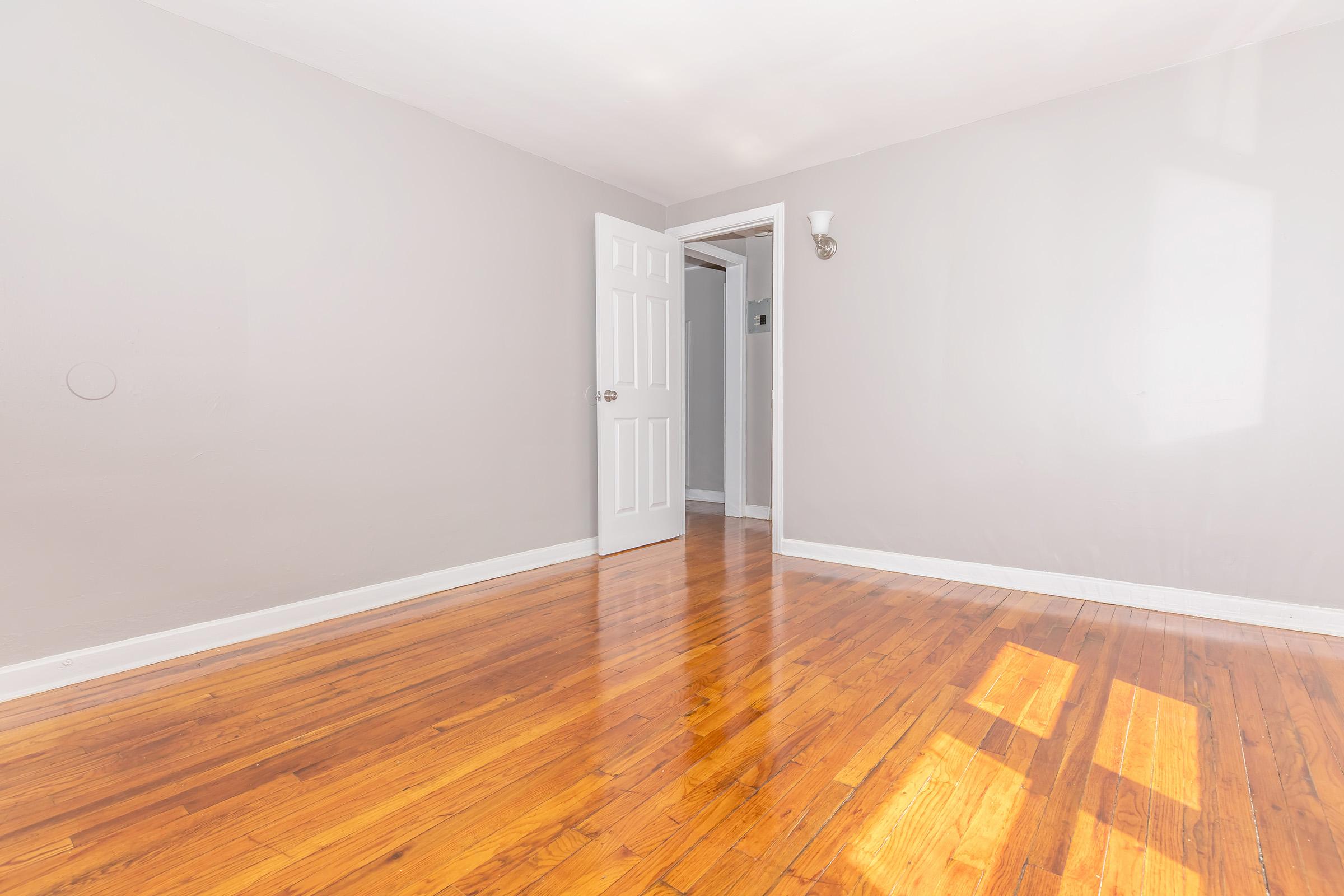 A spacious, empty room with light gray walls and polished hardwood flooring. A white door is open, leading to another area, and natural light streams in, creating soft shadows on the floor.