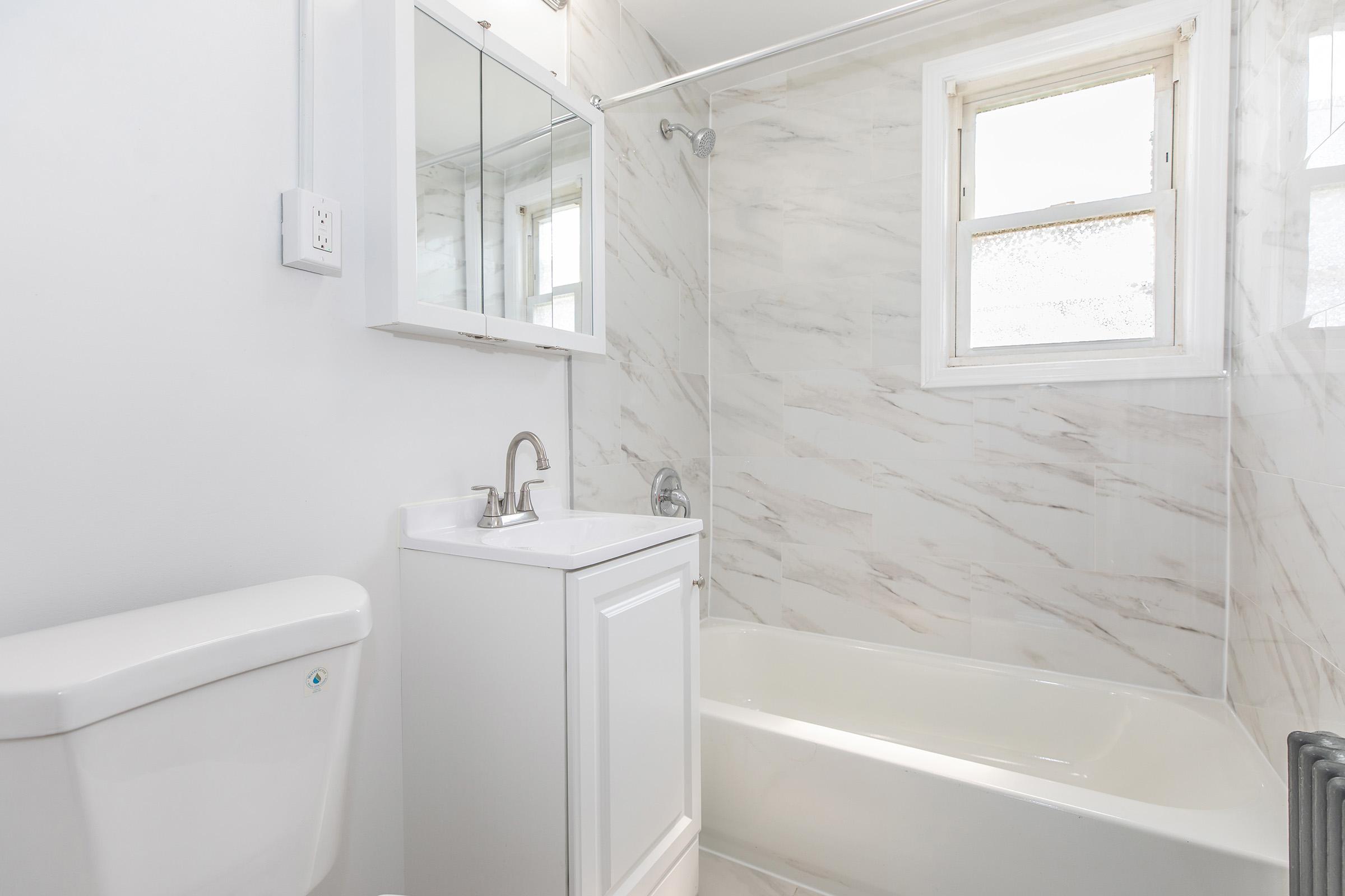 A clean, modern bathroom featuring a white toilet, a sleek vanity with a sink, and a bathtub. The walls are decorated with light marble tiles, and there is a large window that lets in natural light. A mirror is mounted above the sink, reflecting the bright, airy space.