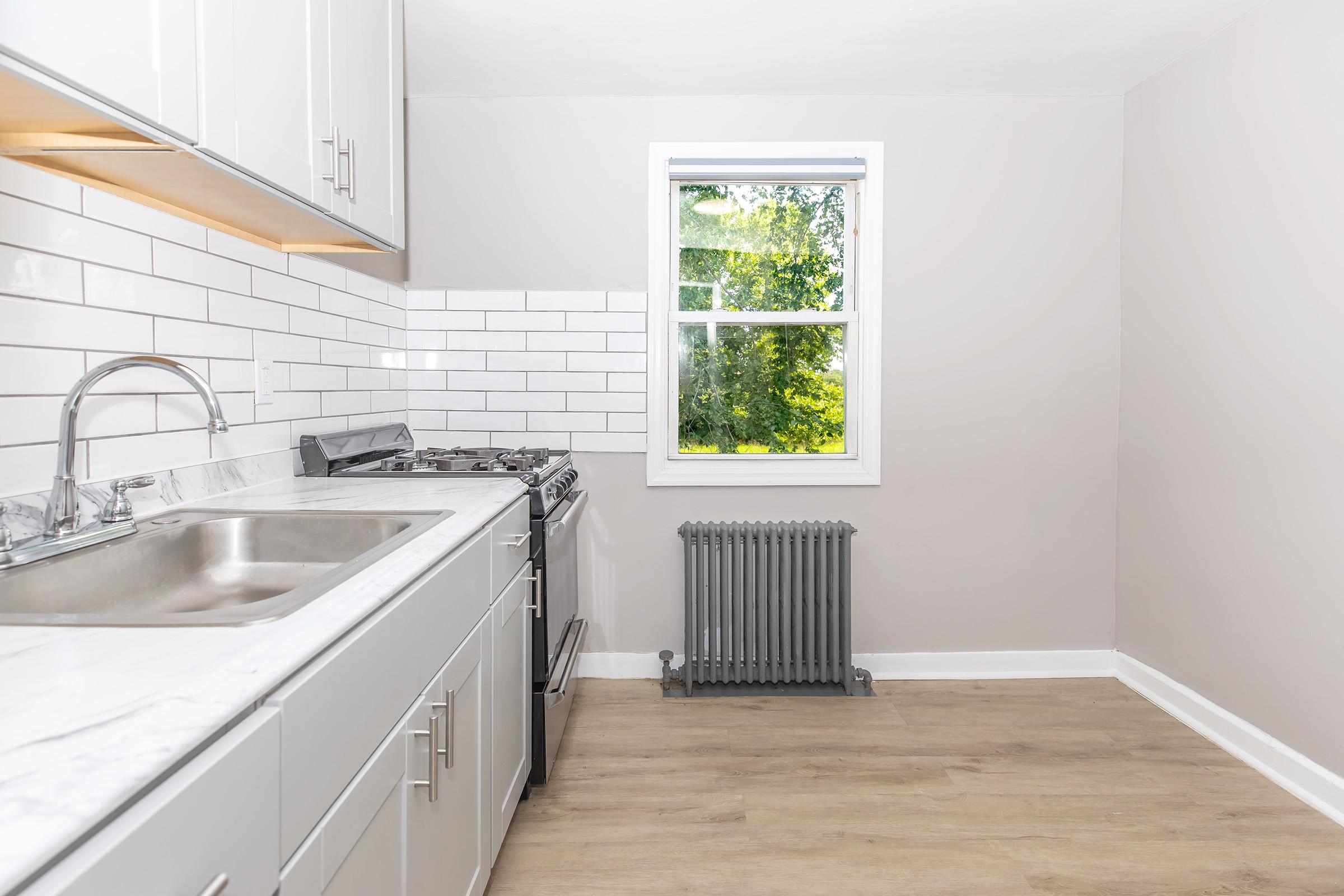 A modern kitchen featuring gray cabinets, a stainless steel sink, and a gas stove. The backsplash consists of white subway tiles, and a window allows natural light to fill the room. A vintage-style radiator is present below the window, and the floor has a light wood finish.
