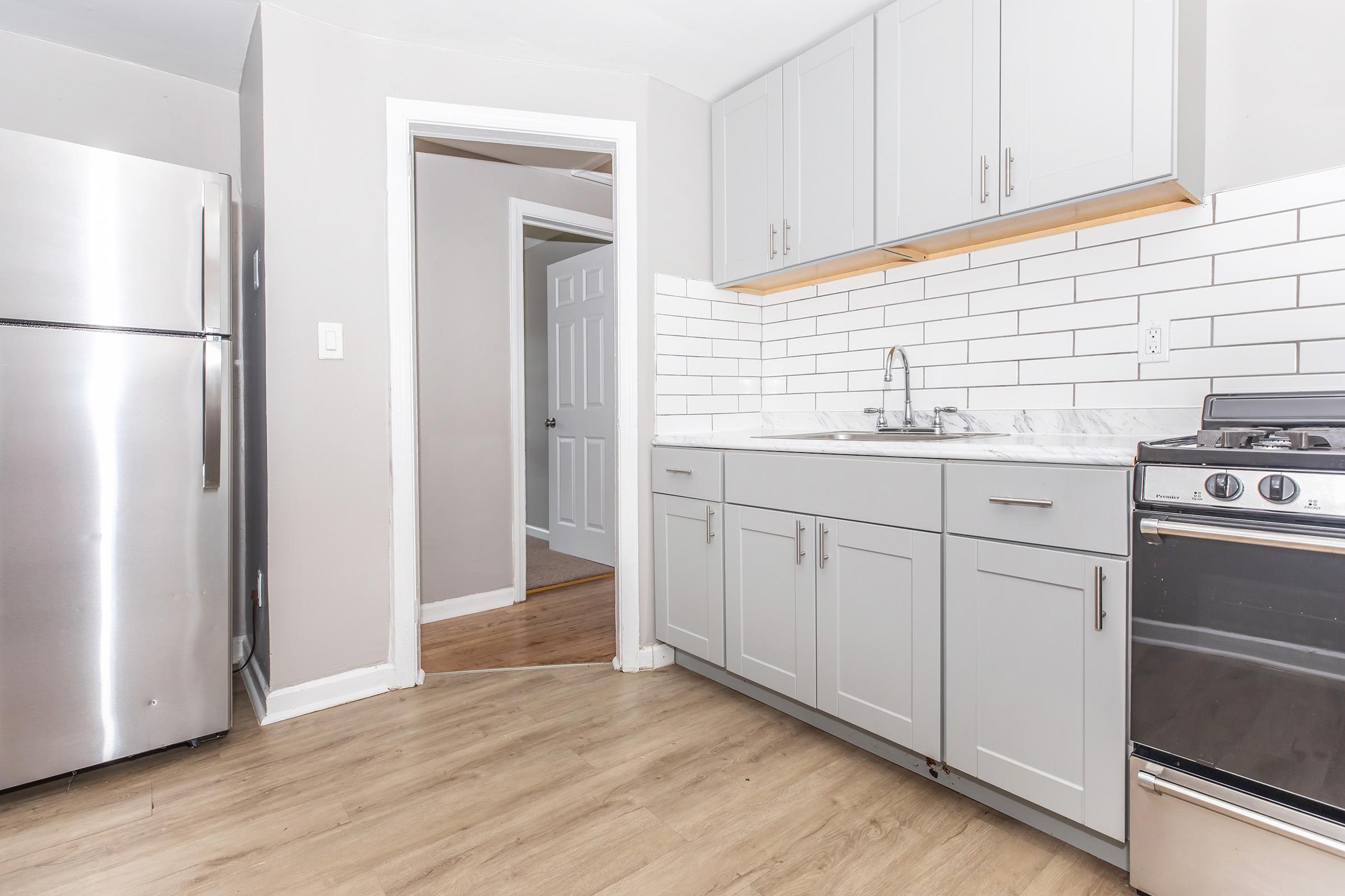 A modern, well-lit kitchen featuring gray cabinets, a stainless steel refrigerator, and a gas stove. The countertop is marble, and there's a white subway tile backsplash. A doorway leads to another room, with wood flooring visible.