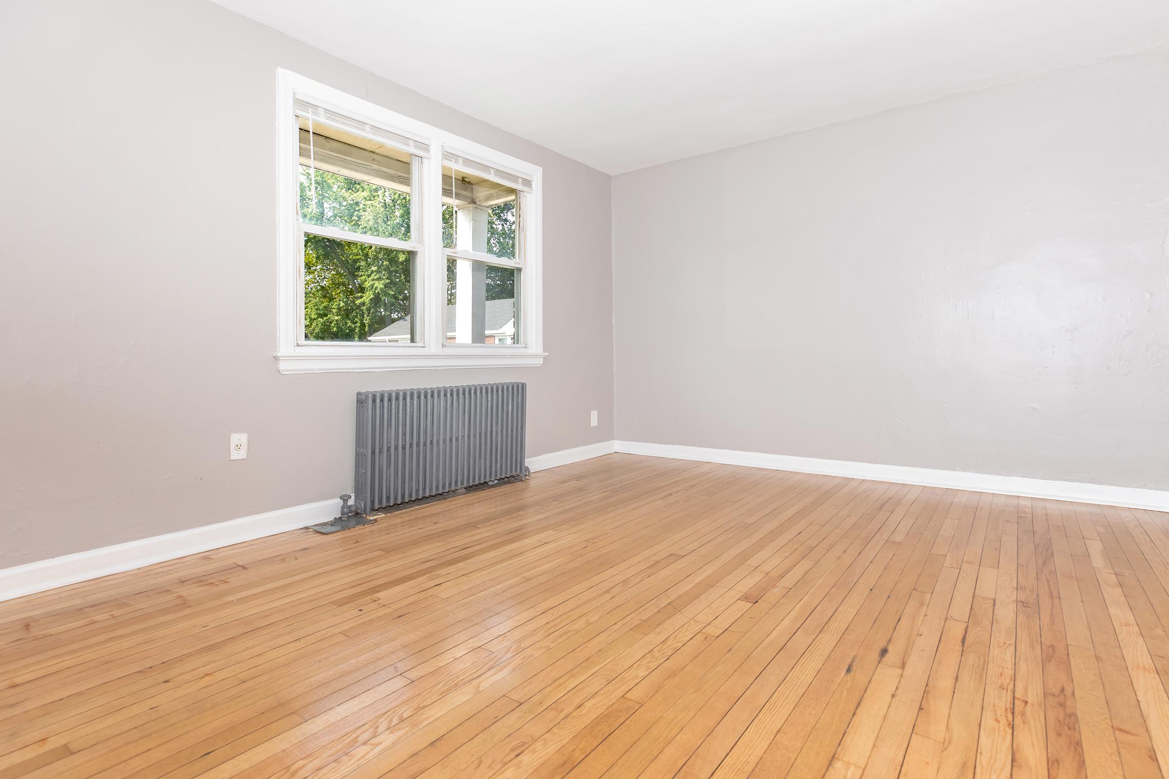 A spacious, empty room with light gray walls and a wooden floor. Two windows let in natural light, and there is a radiator beneath one window. The overall atmosphere is bright and airy, perfect for a new design or furniture arrangement.