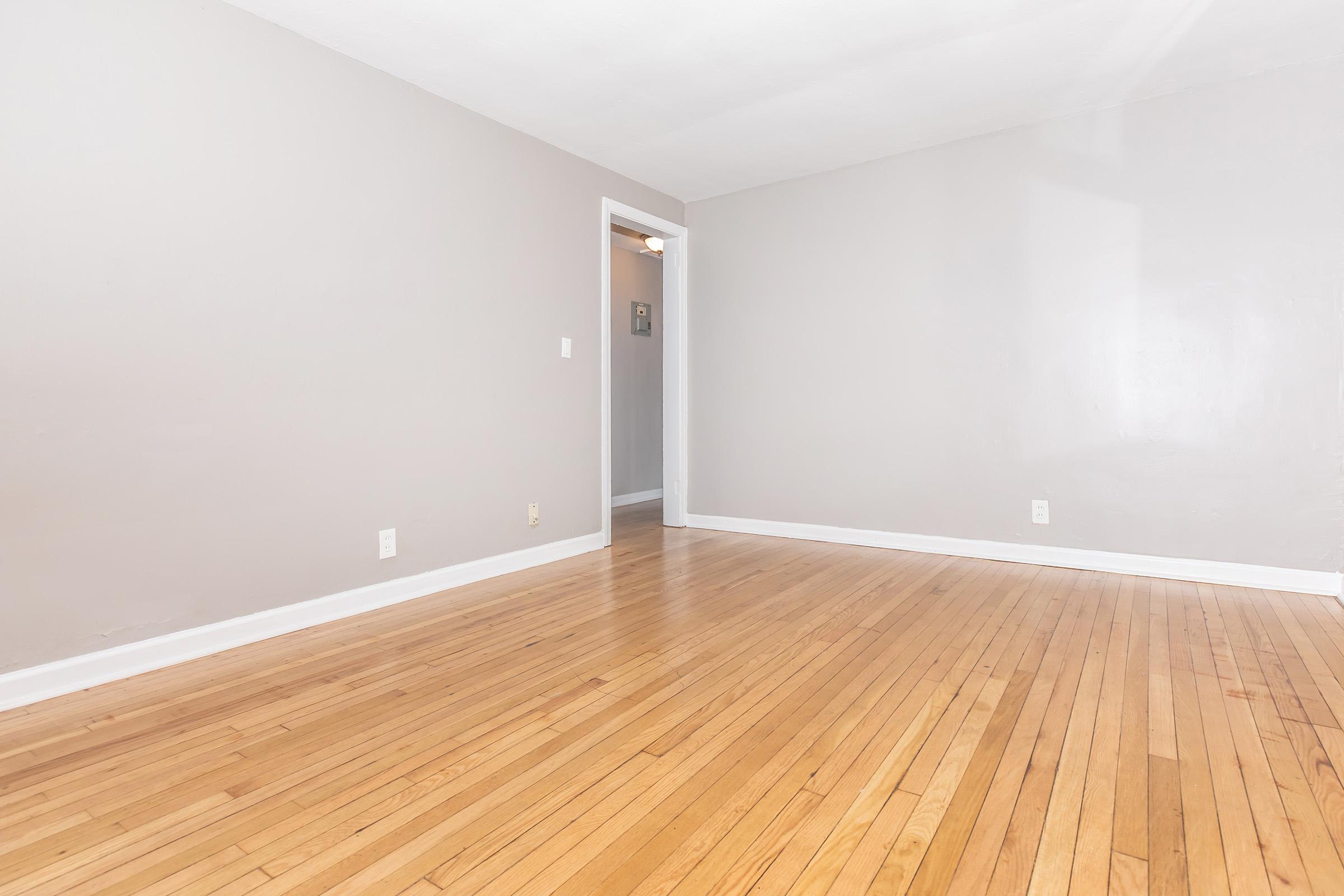 A spacious, empty room with light gray walls and hardwood flooring. The room has a white door leading to another space, and bright natural light illuminates the area from an unseen window. The overall atmosphere is clean and minimalistic.