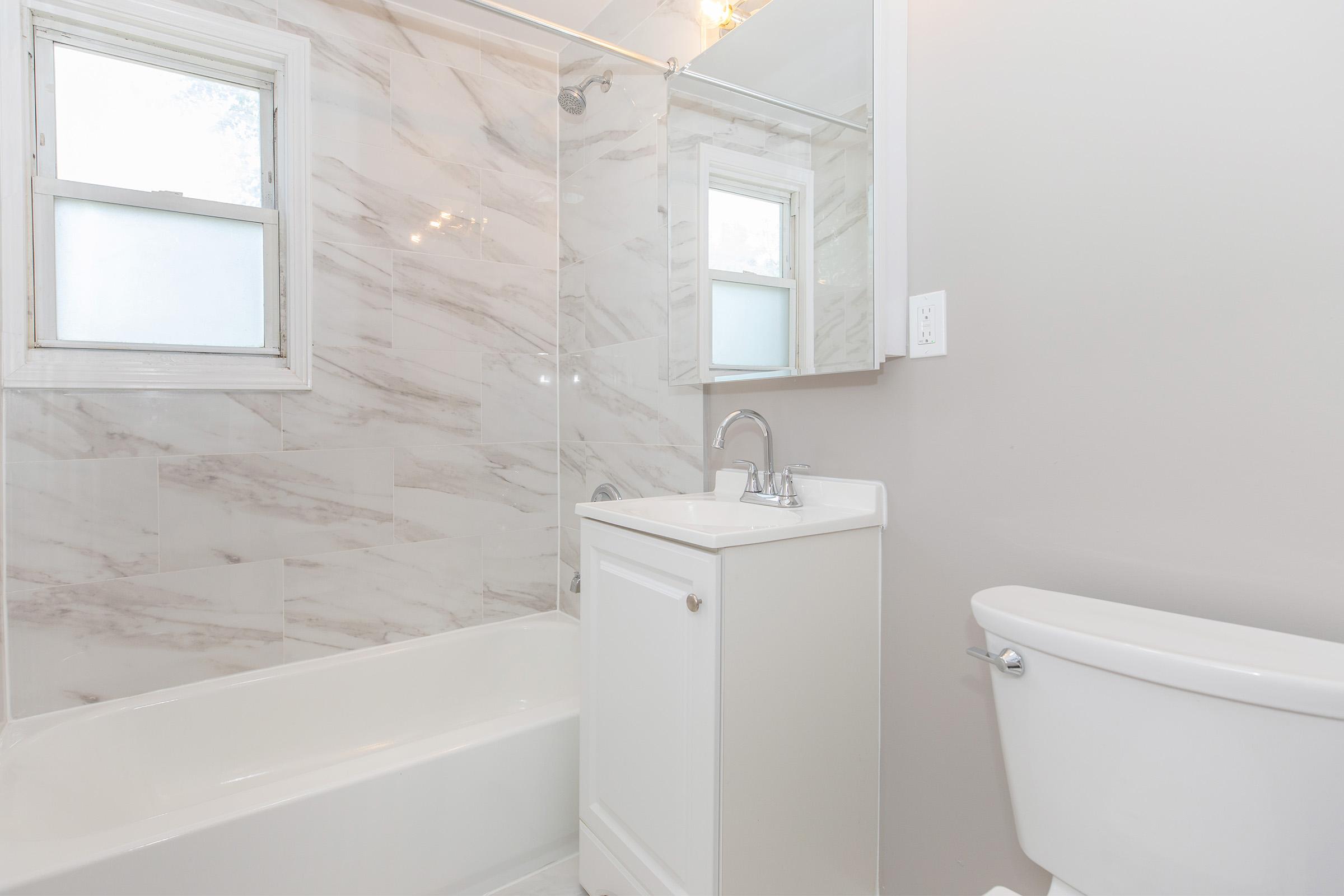 A clean and modern bathroom featuring a white bathtub, a glass shower enclosure, a compact vanity with a sink, and a wall-mounted mirror. The walls are adorned with light marble tiles, while the floor is white. There is a white toilet next to the vanity. Natural light enters through a small window.