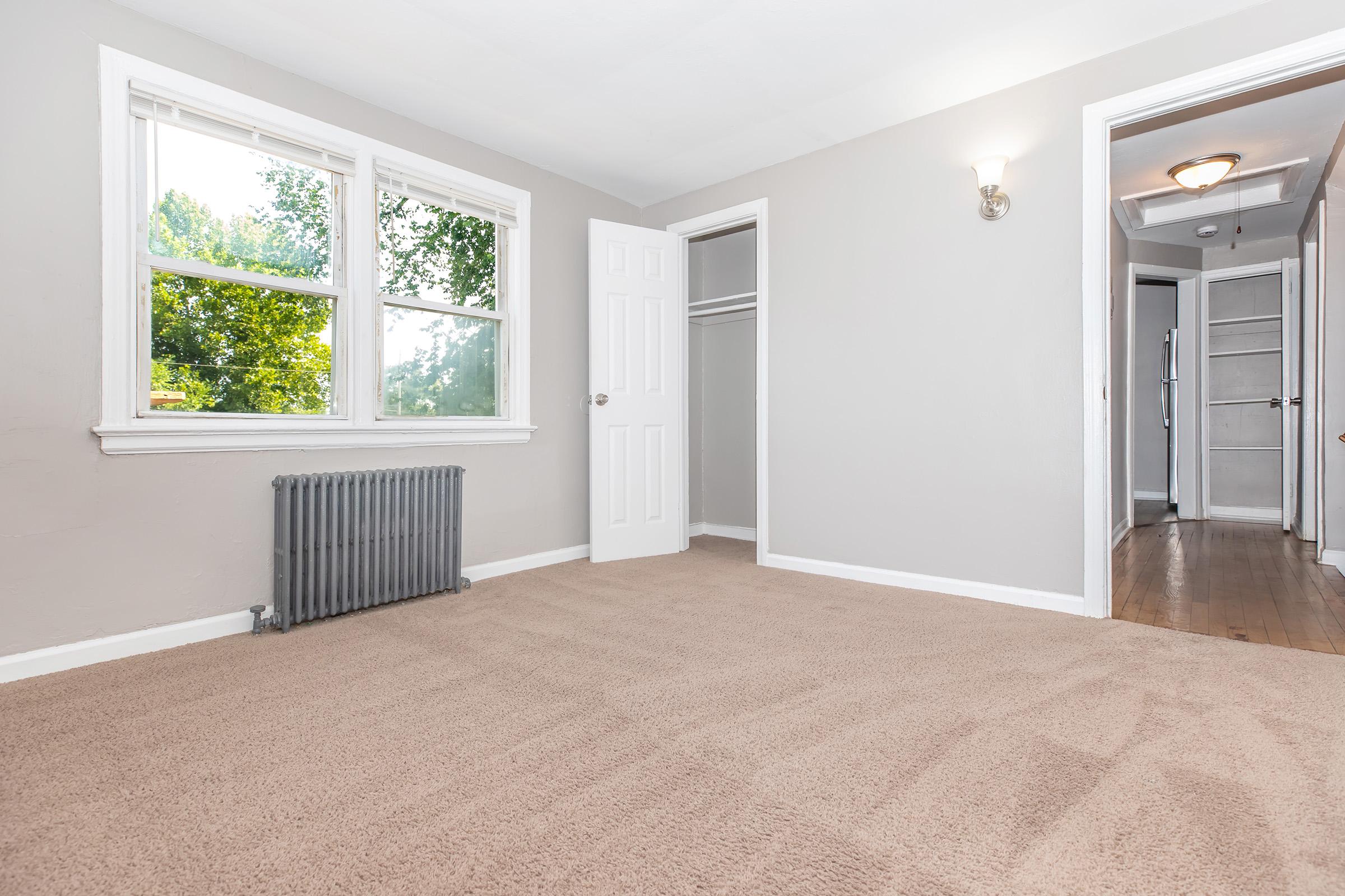 Empty room with light gray walls and beige carpet. Two large windows let in natural light, showing green trees outside. There is a white door leading to a closet and another door that opens to a hallway. A vintage radiator is located on the left wall, adding charm to the space.