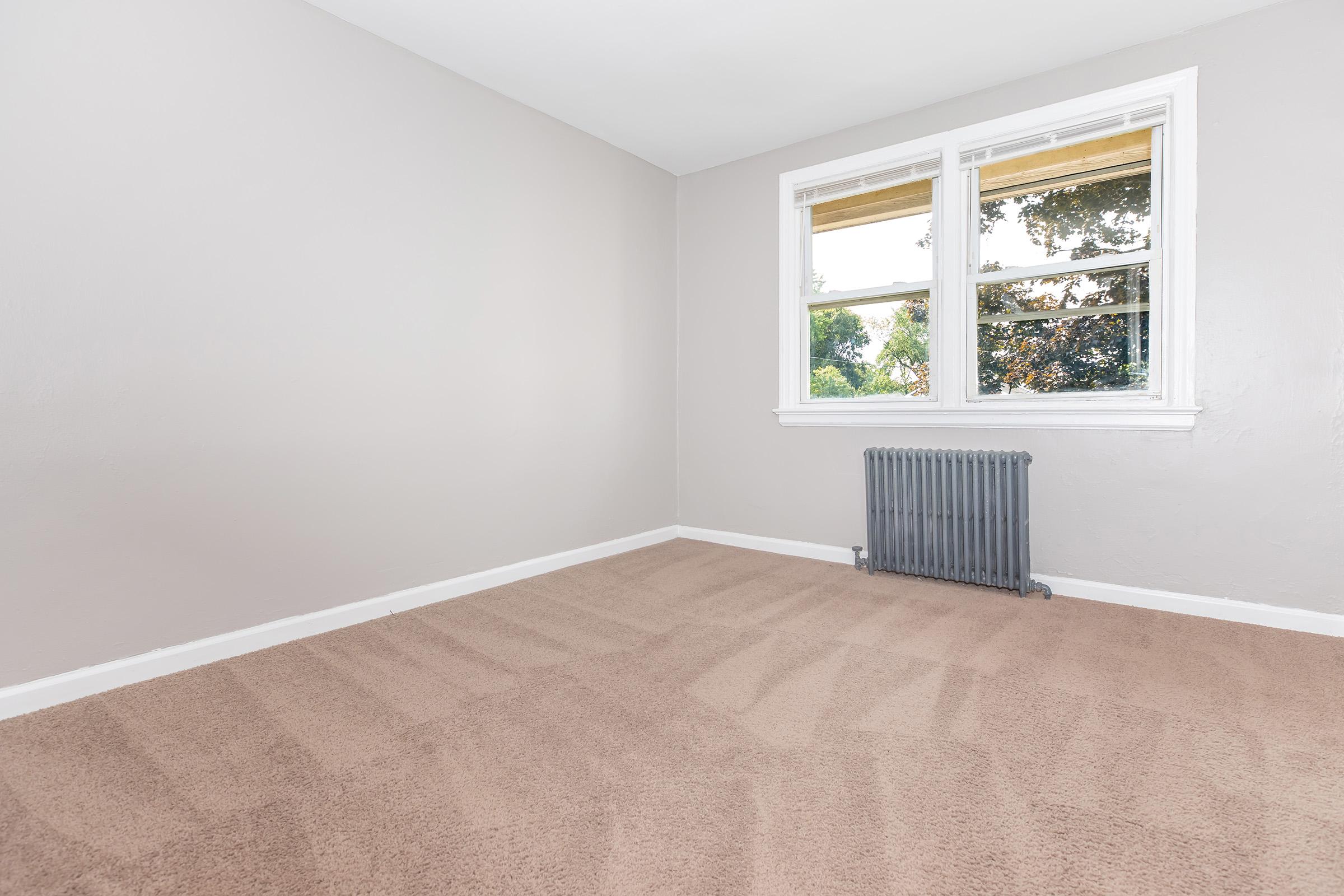 Empty room with light gray walls and a tan carpet. It features two windows with beige shades allowing natural light, and a small radiator beneath the windows. The room is uncluttered, creating a spacious feel.