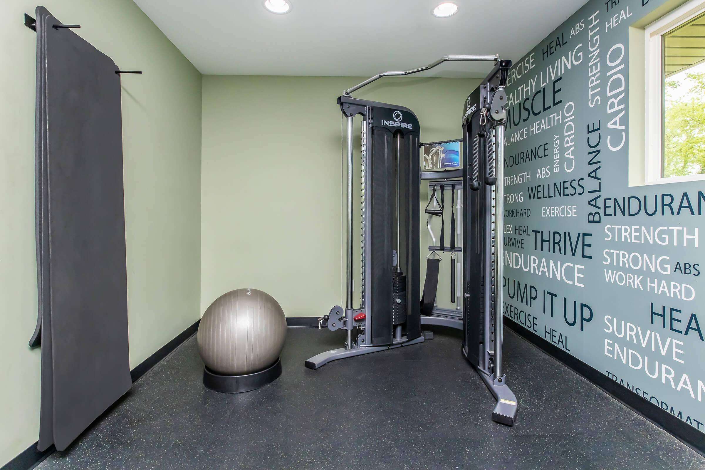 A modern fitness room featuring a multi-purpose gym machine, an exercise ball, and a wall adorned with motivational phrases related to health and wellness. The space is well-lit with natural light and has an overall clean, organized appearance, ideal for workouts and training.